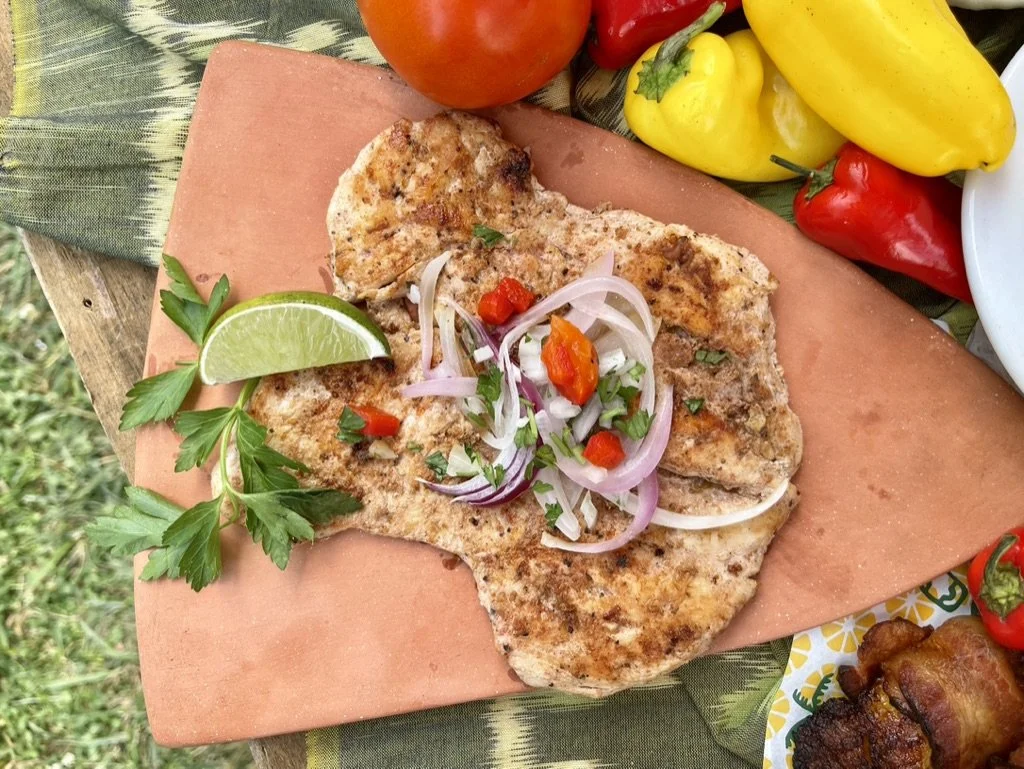 Grilled meat topped with sliced onions, red chili peppers, and herbs, garnished with a lime wedge and parsley, on a pink stone surface, with colorful peppers and vegetables in the background.