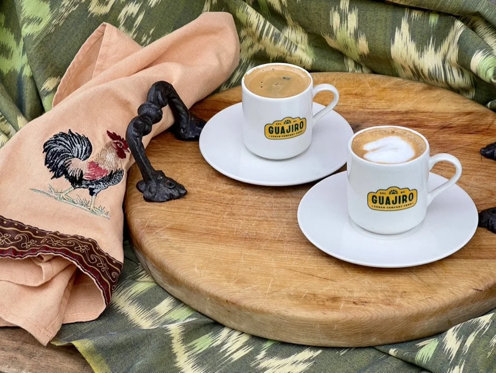 Two cups of coffee on white saucers placed on a round wooden table. One coffee has foam art, and the other appears to be black coffee. A beige cloth with a rooster embroidery and red and brown accents, along with dark decorative figurines, are on the