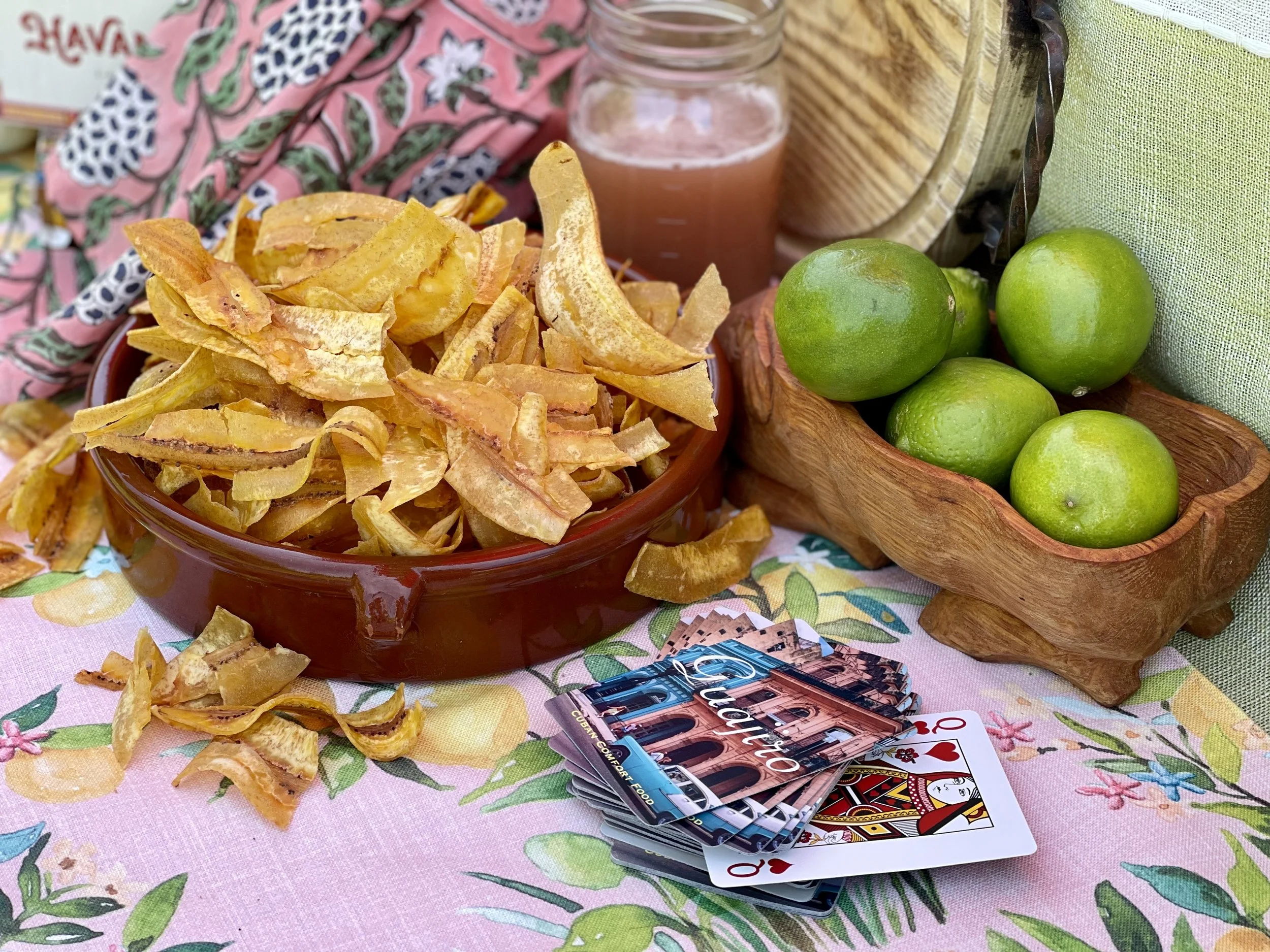 A table with dried fruit chips in a bowl, green limes in a wooden basket, a glass of pink juice, colorful brochures, and a deck of playing cards, all arranged on a floral tablecloth.