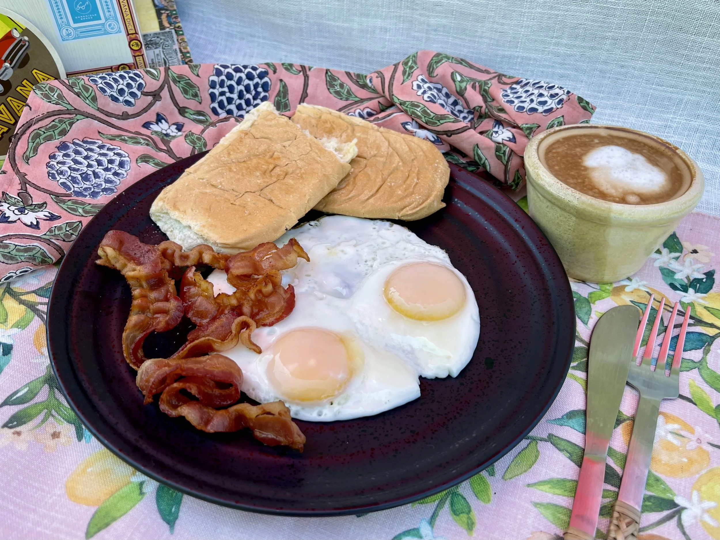A breakfast plate with two fried eggs, crispy bacon, toasted bread, and a cup of coffee on a colorful floral tablecloth.
