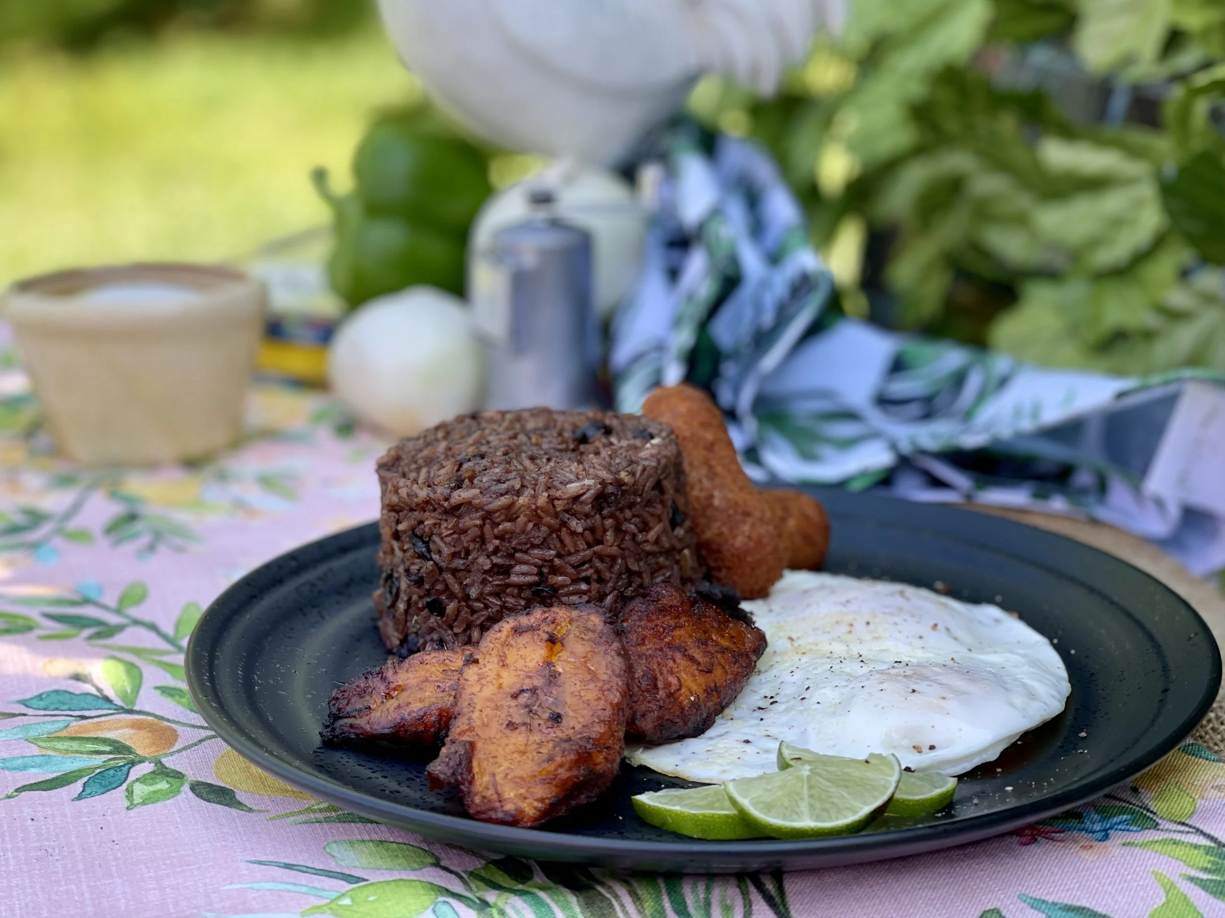 A plate of Cuban breakfast food including chorizo and eggs, a slice of avocado toast, lime slices, and rice, set on a table with a floral tablecloth.