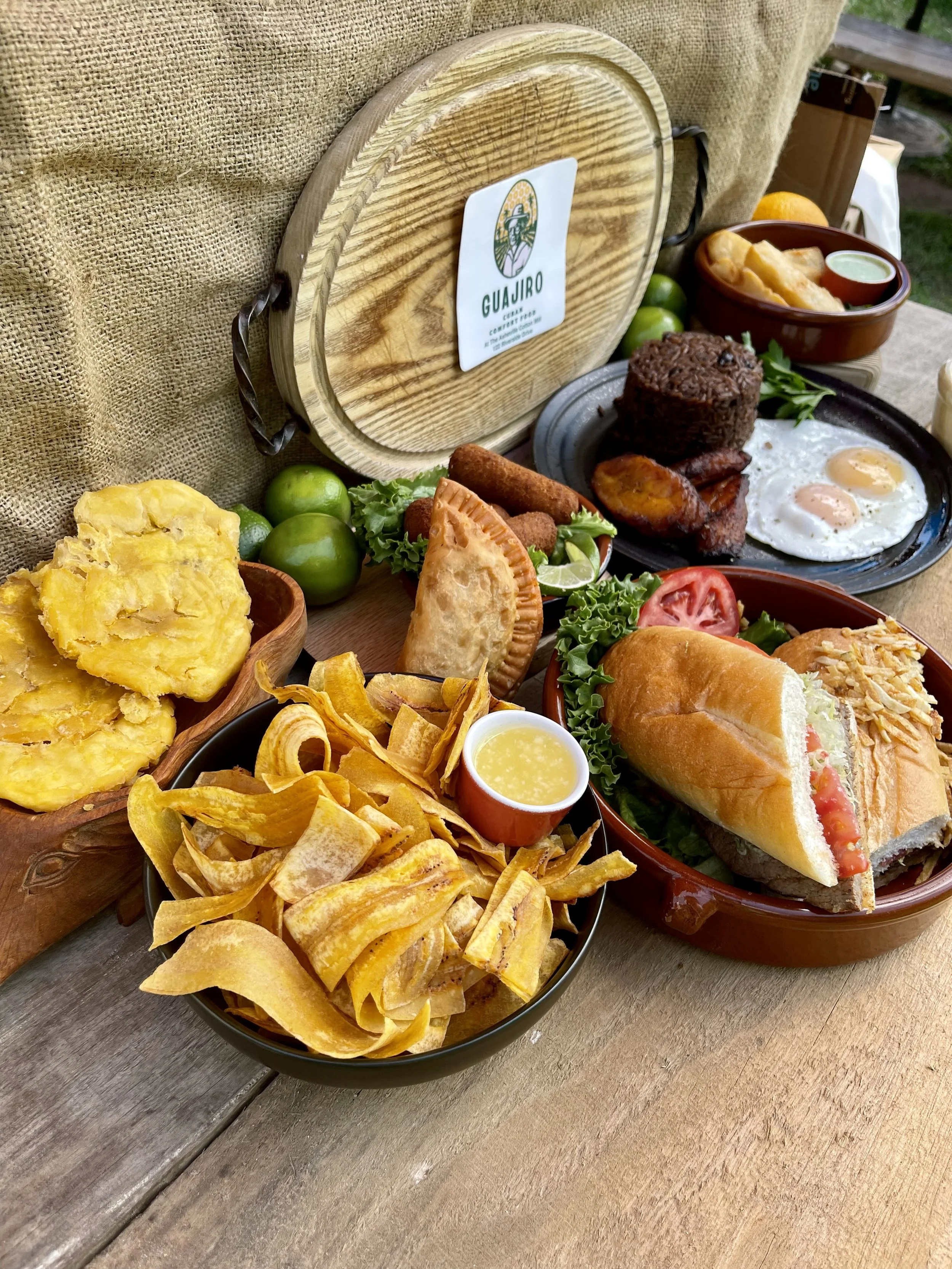 Platter of traditional Cuban breakfast foods including eggs, fried plantains, nachos, a sandwich with tomato and lettuce, and various side dishes on a wooden table.