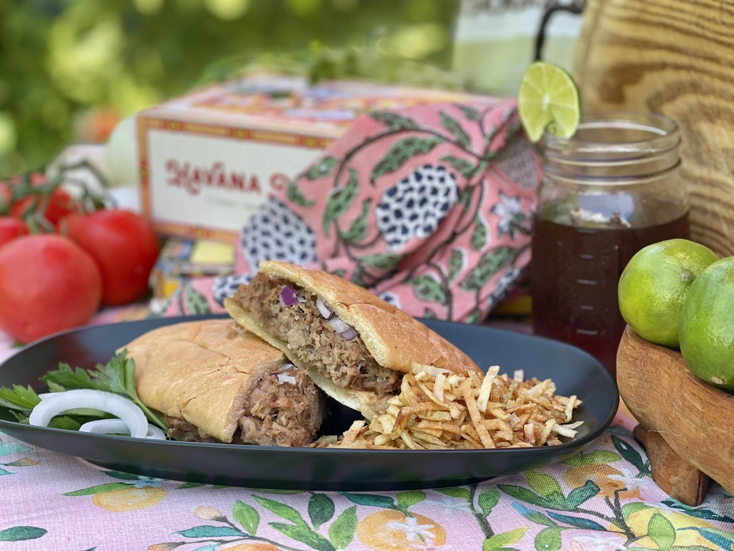 A plate with a beef taco topped with chopped onions and cilantro, with shredded cheese on the side. Behind the plate, there is a jar of dark beverage with a lime wedge, a box of La Victoria enchilada sauce, and fresh tomatoes and green grapes. The sc
