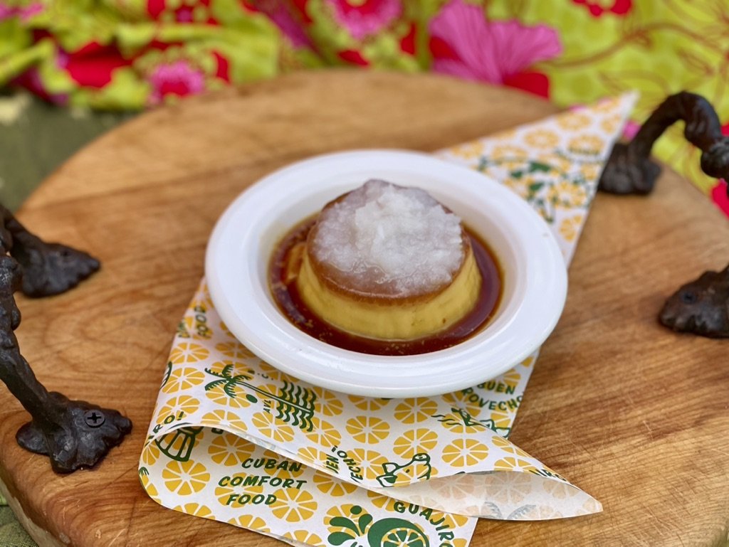 A serving of flan with caramel sauce and a white topping, on a small white plate set on a decorative paper with yellow flowers and green writing, on a wooden cutting board with metal animal legs.