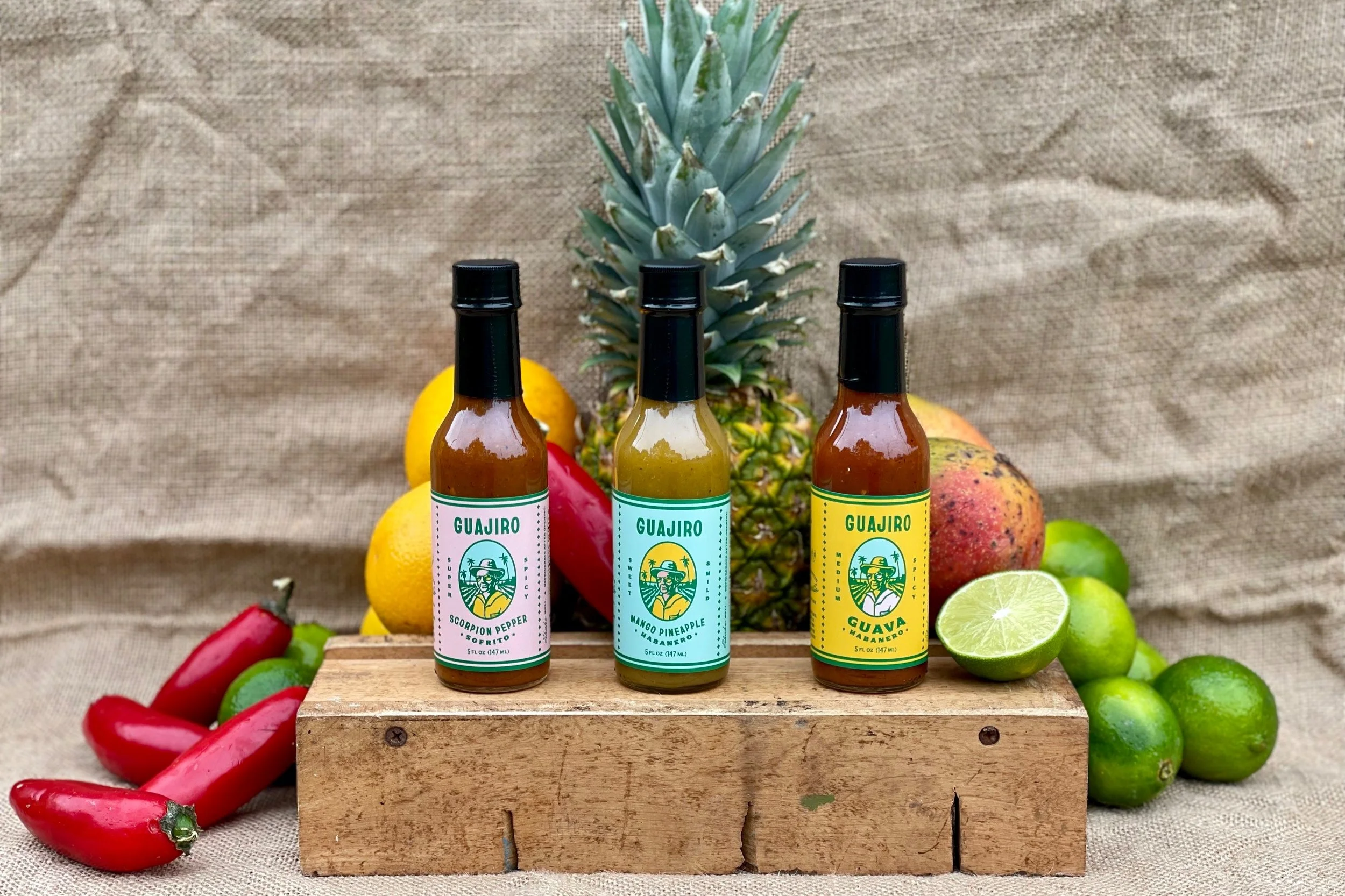 Three bottles of Guajiro crispy pepper hot sauces, with a pineapple, apples, lemons, and red chili peppers surrounding a wooden box, against a beige fabric background.