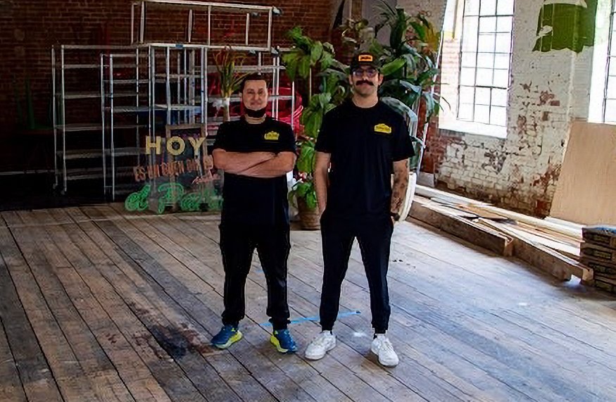 Two men in black shirts and pants standing in a rustic indoor space with exposed brick walls, large windows, and wooden flooring, with some greenery and signage in the background.
