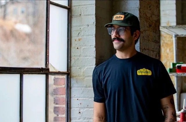 A man with glasses, a mustache, and a beard, wearing a navy T-shirt and a cap, standing near a window and looking outside in an industrial-style room.