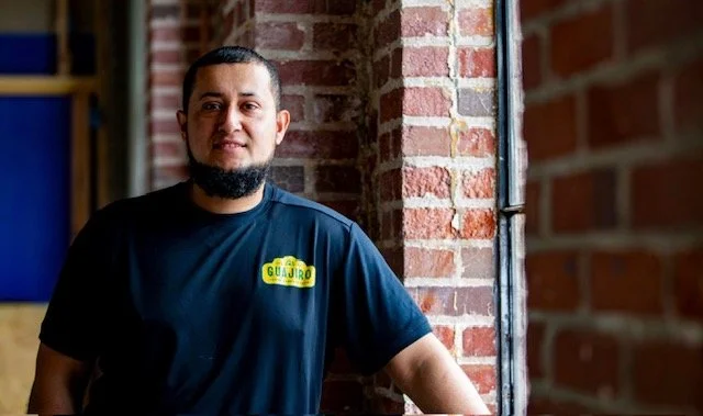 A man with a beard and short hair wearing a black shirt with a yellow logo, standing next to a brick wall and window inside a building.
