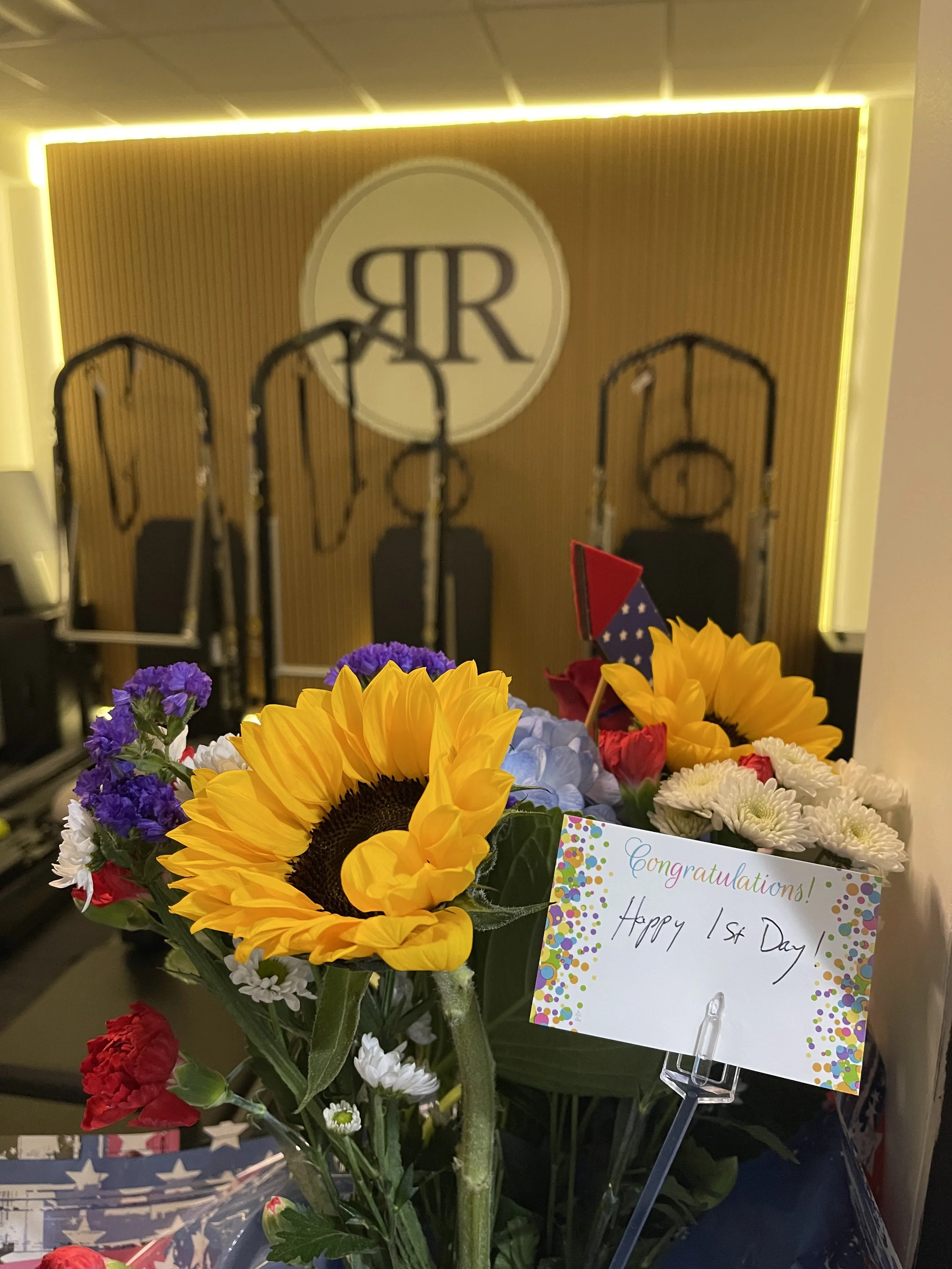 A bouquet of colorful flowers, including sunflowers, red, blue, purple, and white blooms, with a congratulatory card that reads 'Congratulations! Happy 1st Day.'