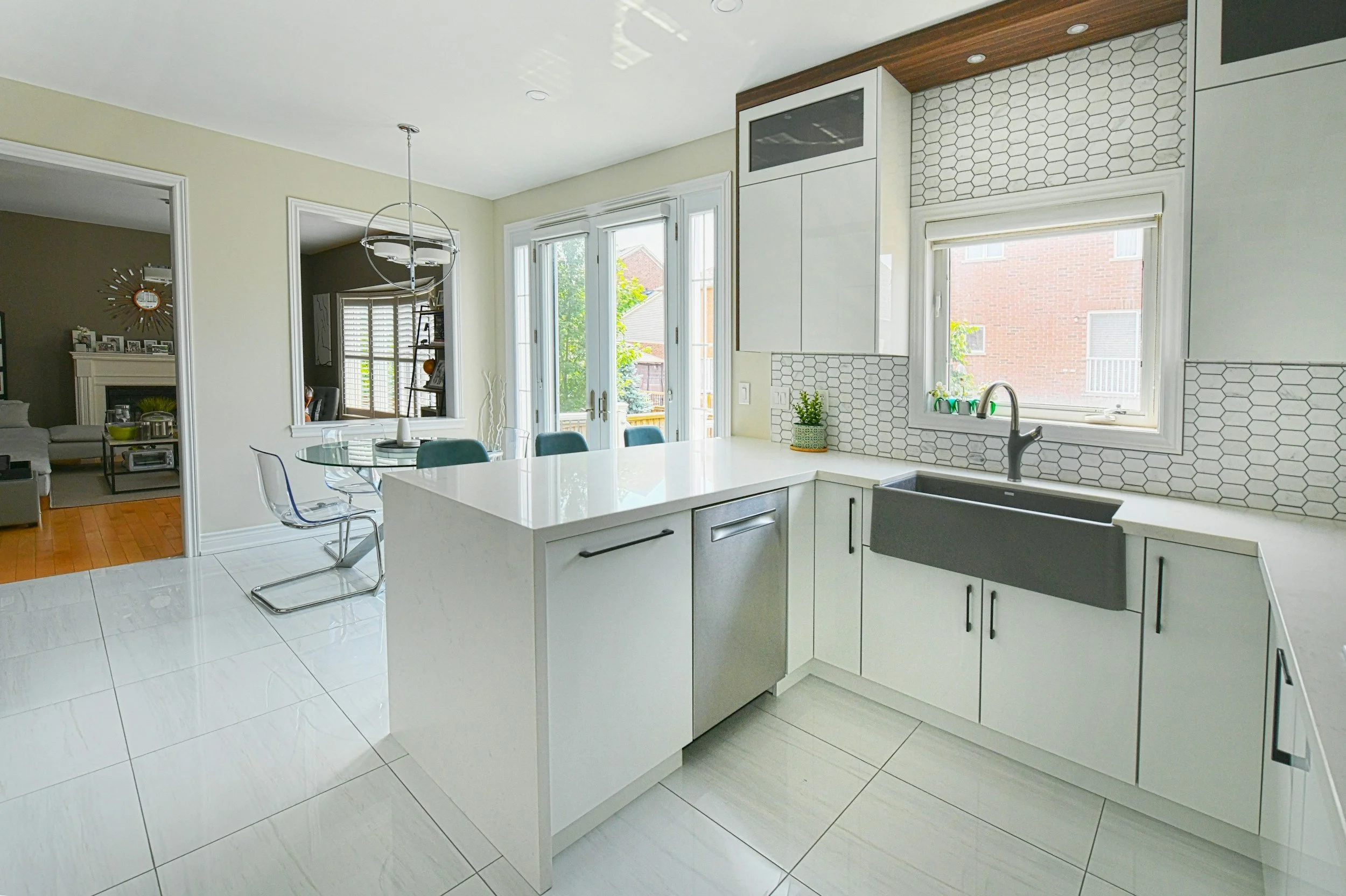 Modern kitchen with white cabinets, gray farmhouse sink, and honeycomb tile backsplash, overlooking a sunlit outdoor area through large windows.