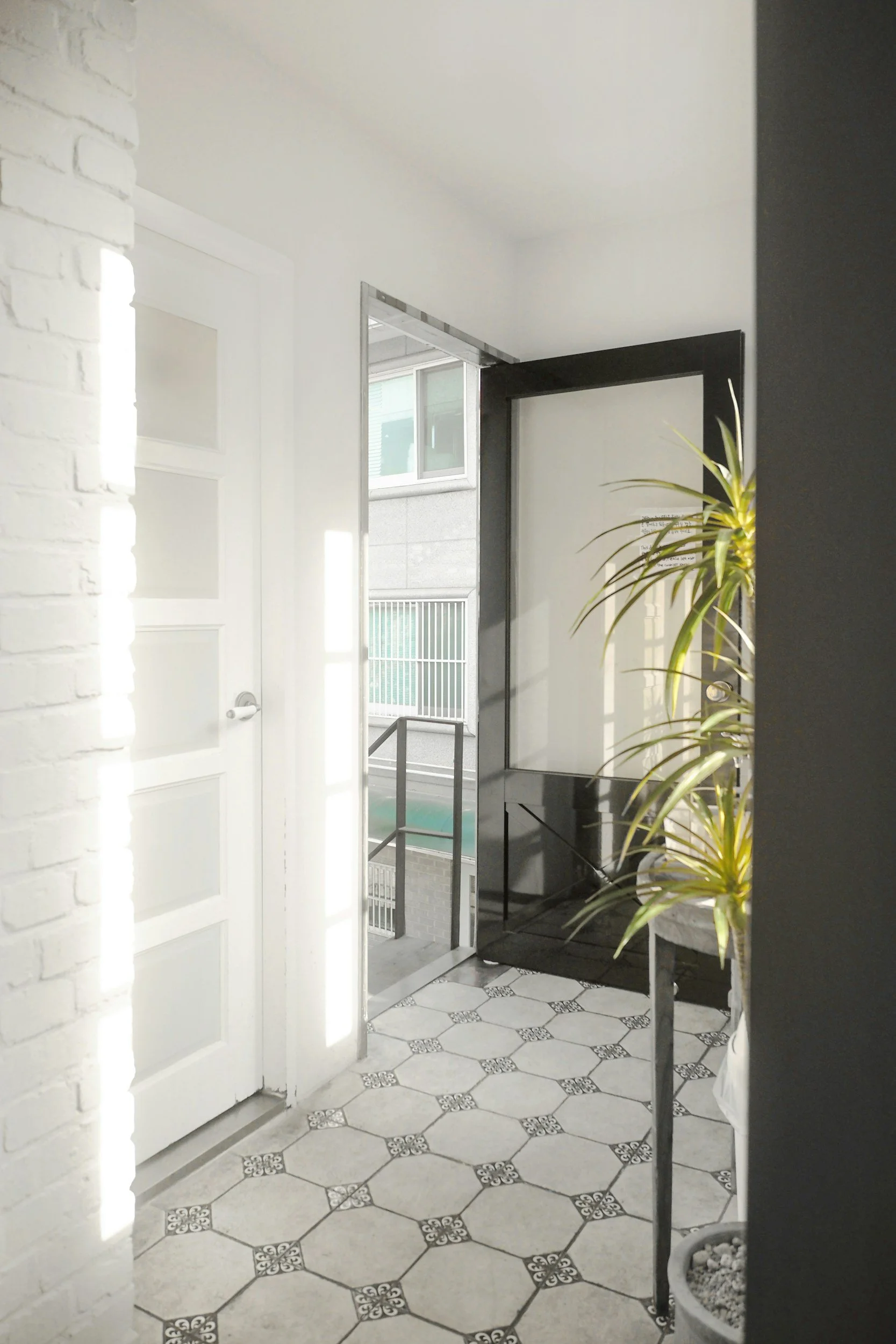 Interior view of a modern building with a tiled floor, white brick wall on the left, and large window with glass doors leading to an outdoor balcony or patio area. There is a black-framed mirror or framed opening and a potted plant on the right.