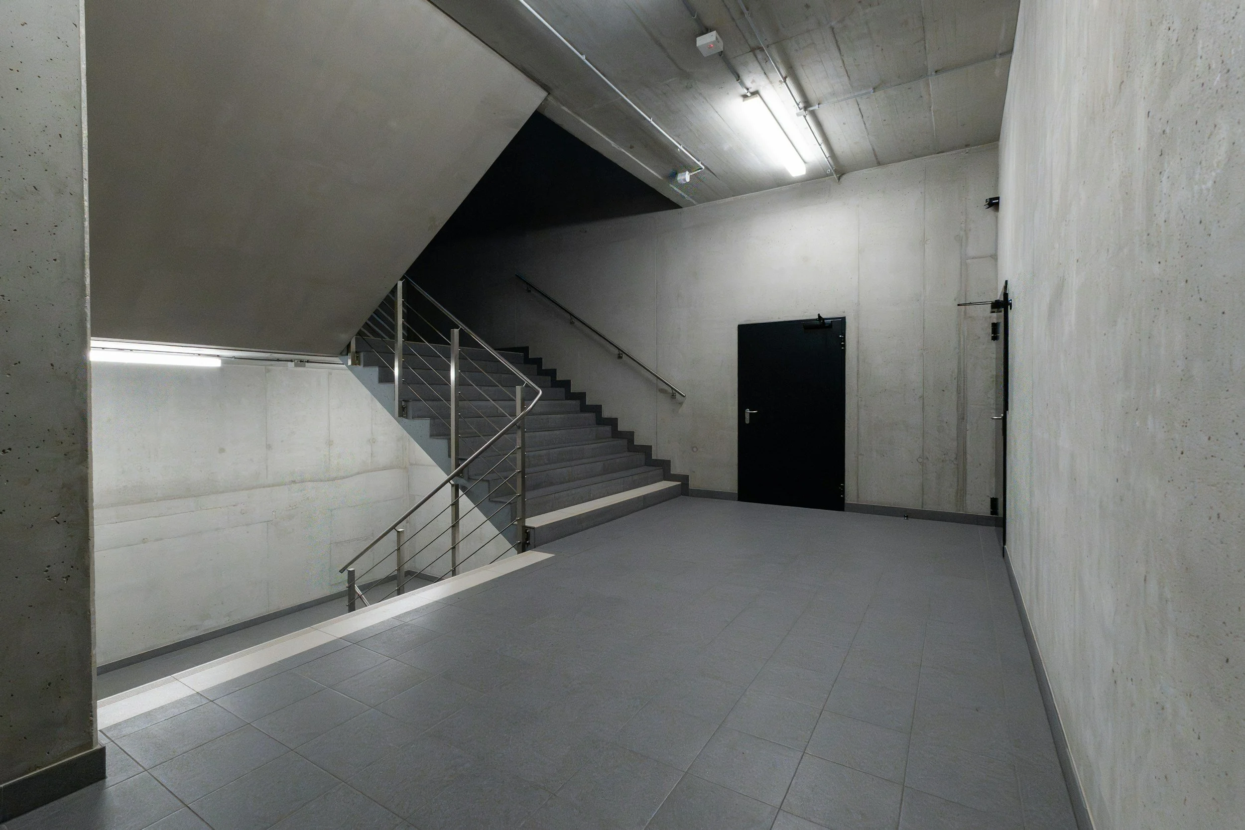 Interior view of a concrete staircase with metal handrails and a black door at the top, illuminated by fluorescent lights on a concrete ceiling.