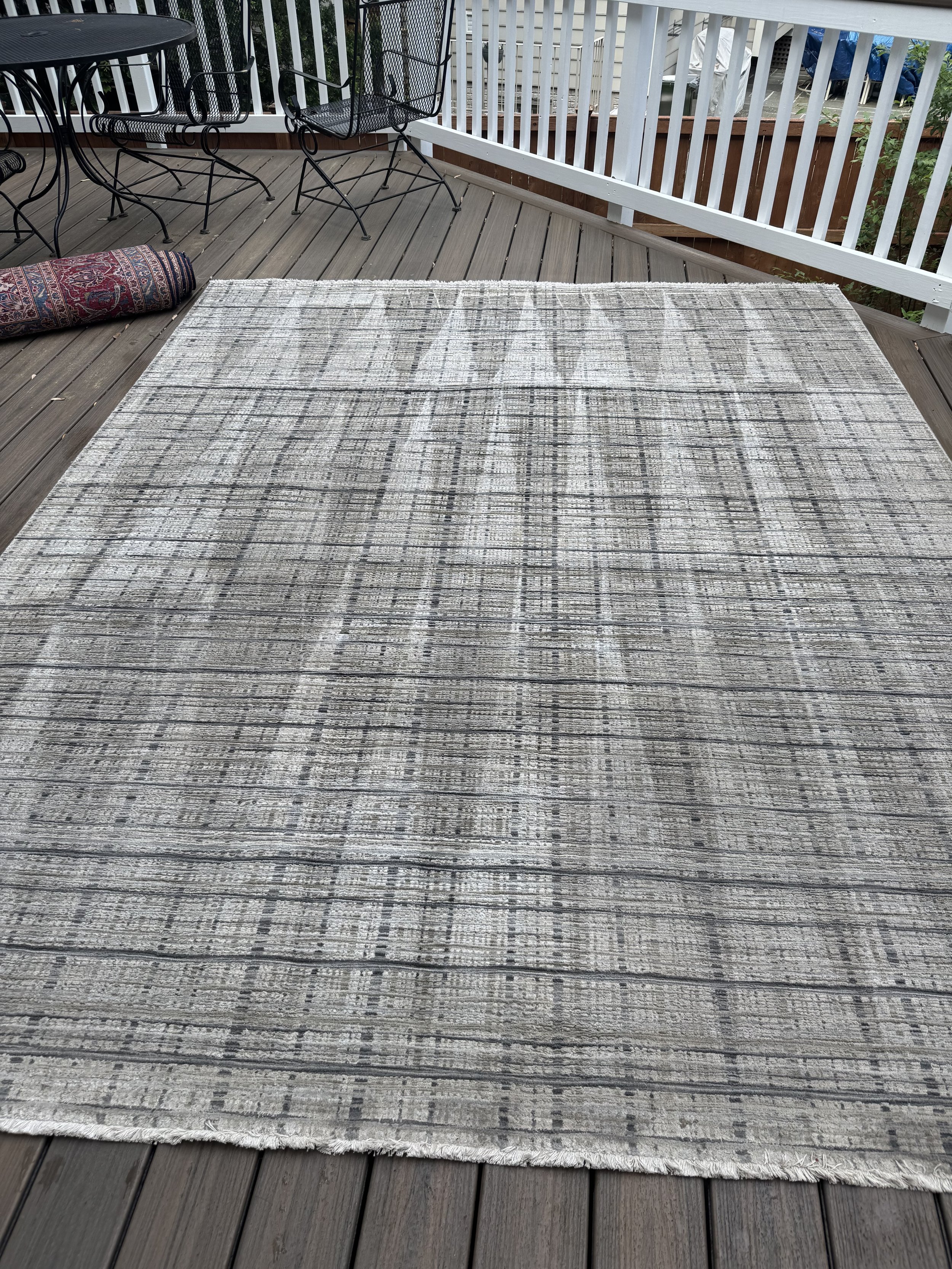 A rectangular area rug with a gray and black striped pattern on a wooden deck, with outdoor furniture and a rolled rug to the side.