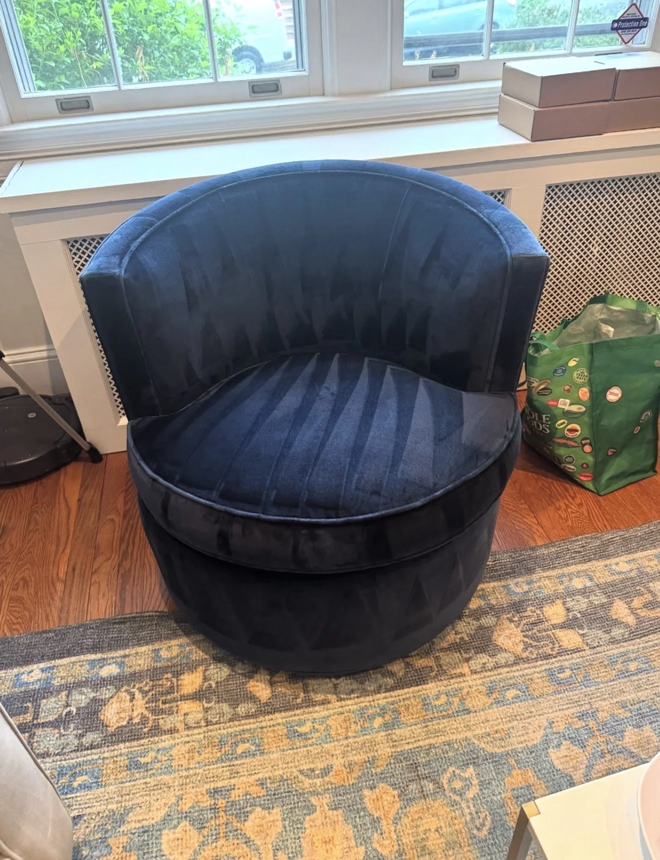 A dark blue, round velvet chair positioned on a patterned area rug in front of a window with white trim. A green shopping bag with colorful stickers sits on the floor to the right of the chair, and a small side table is partially visible in the bottom right corner.