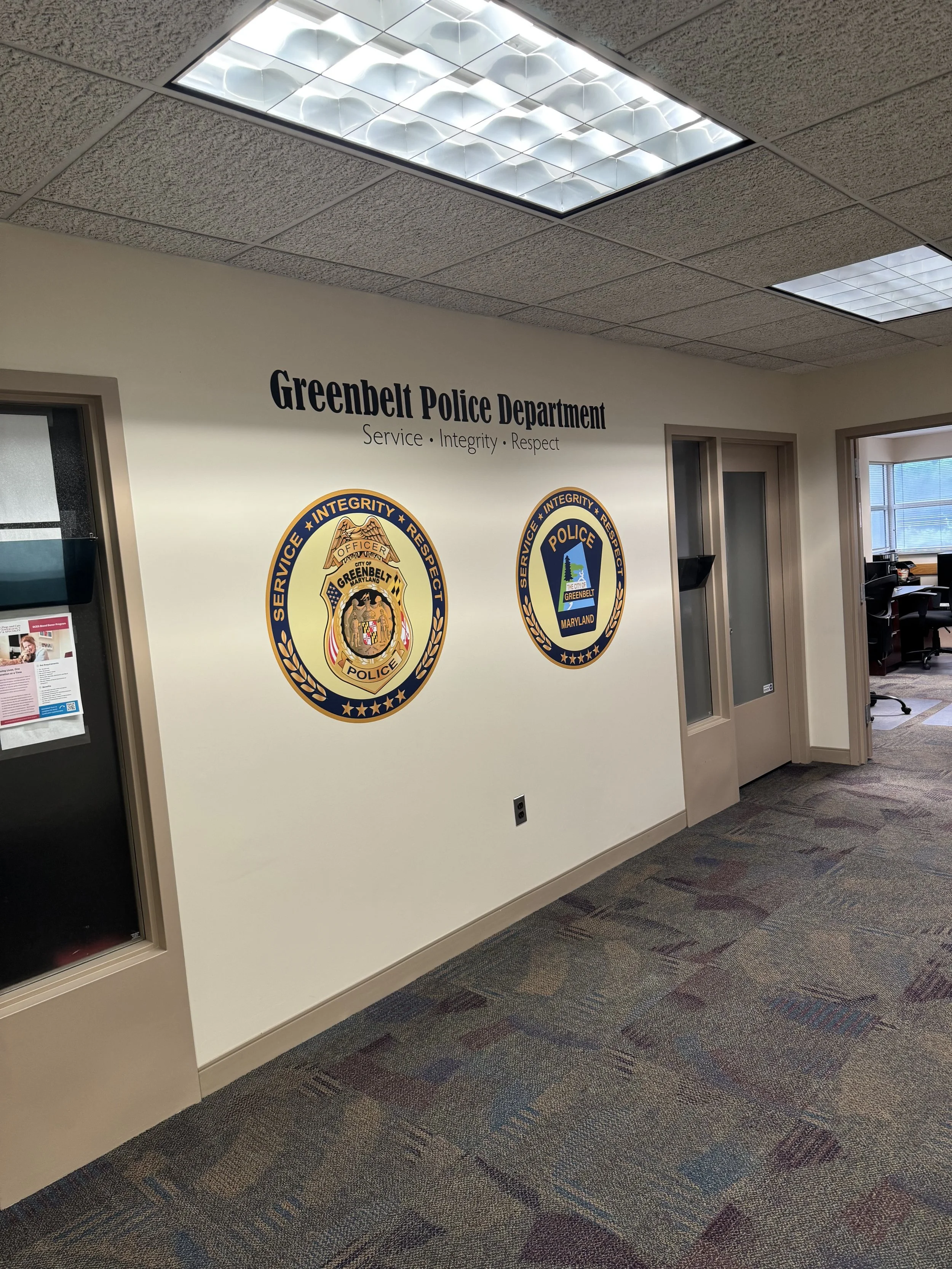 Interior view of the Greenbelt Police Department, showing a wall with the department's name and badges, and part of an office area with chairs and desks.