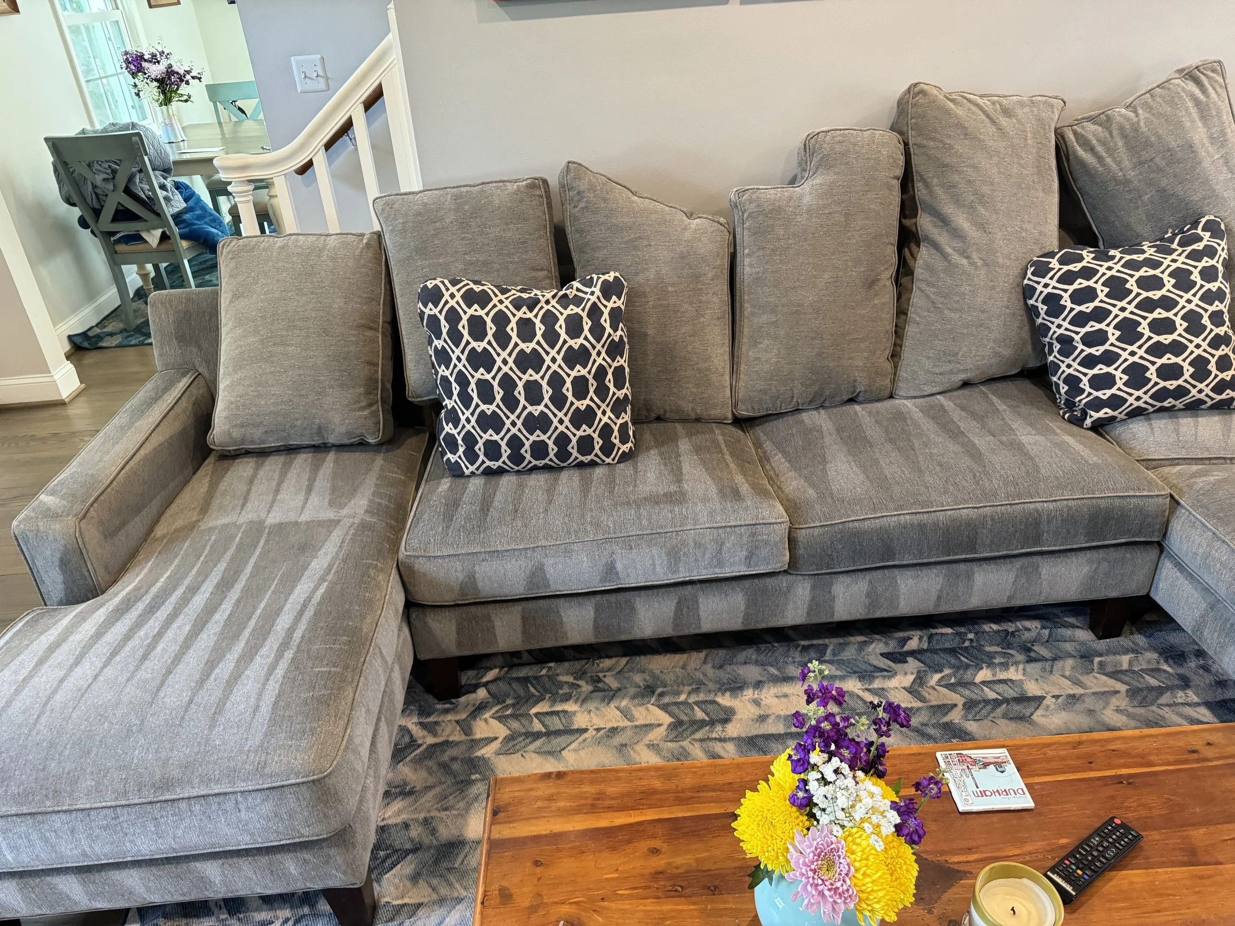 A living room with a grey couch with throw pillows, a wooden table with a colorful bouquet of flowers, and a corner with a dining area visible in the background.
