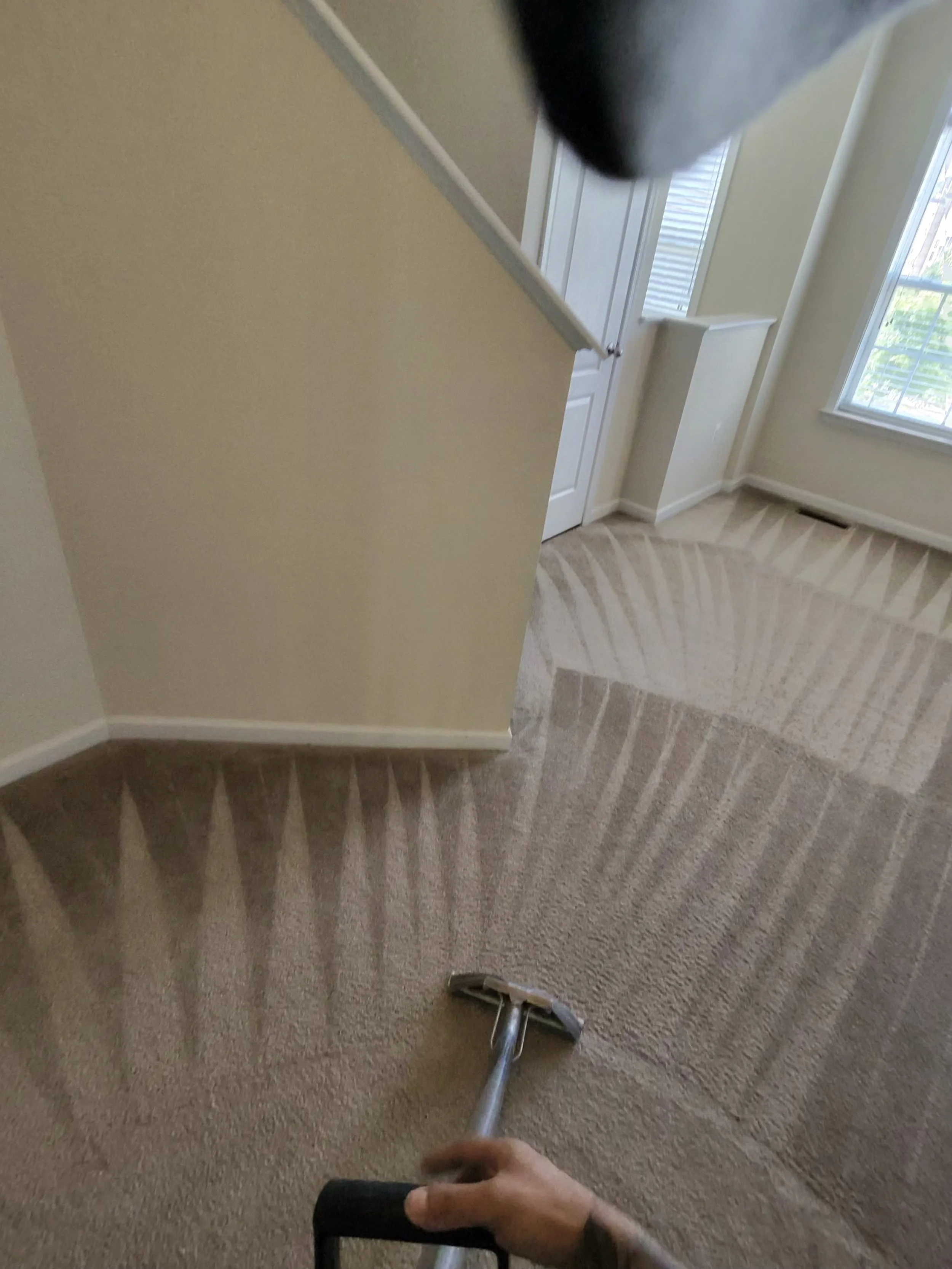 A person vacuuming beige carpet in a house with beige walls, white trim, a window with blinds, and white doors.