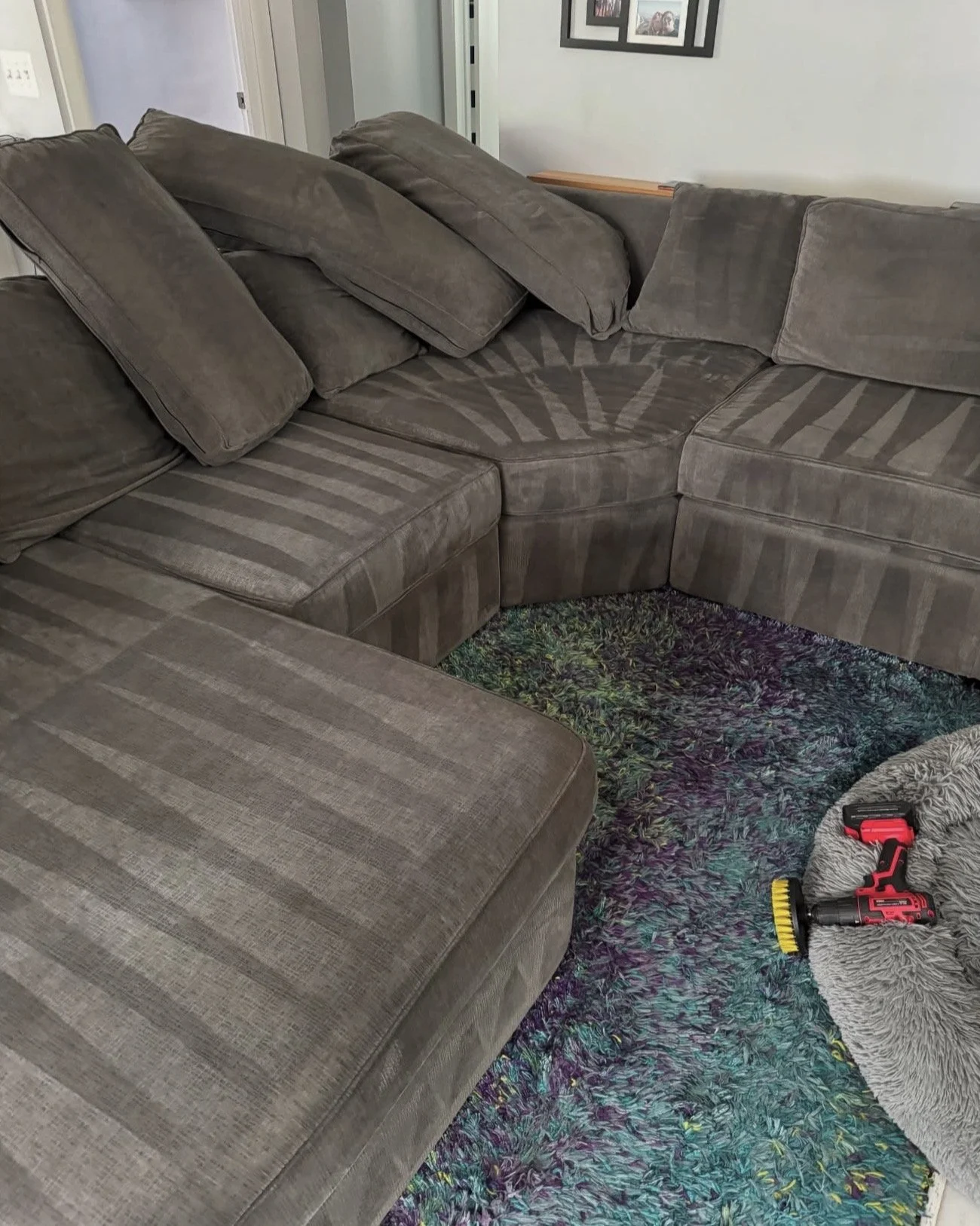 A beige sectional sofa with patterned upholstery and several loose cushions. A colorful rug is on the floor, and tools are placed on a small blanket nearby. Wall with picture frames in the background.