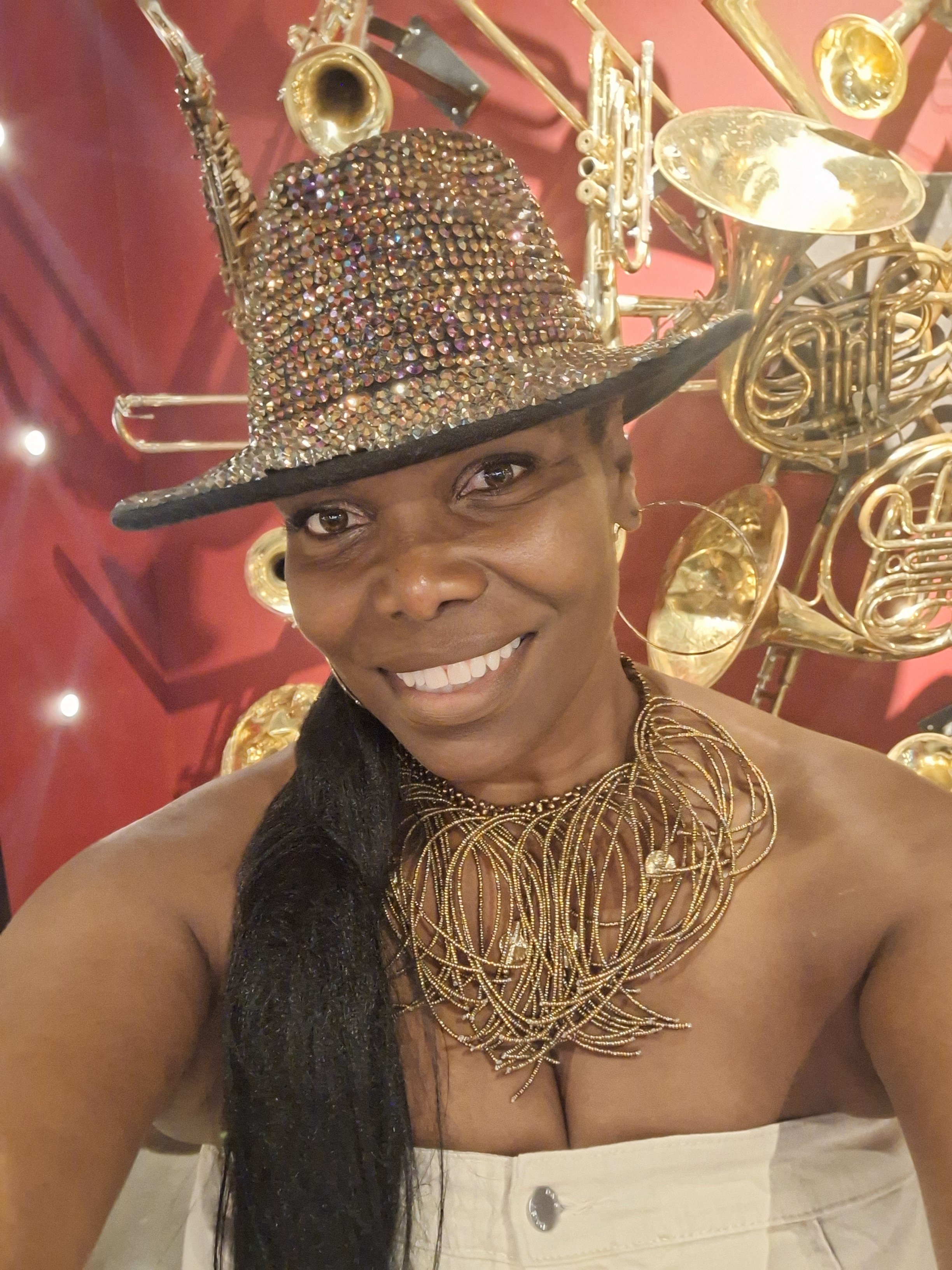 A woman wearing a shiny, rhinestone-studded cowboy hat and elaborate gold jewelry, smiling for a selfie in front of a display of gold-colored musical instruments.