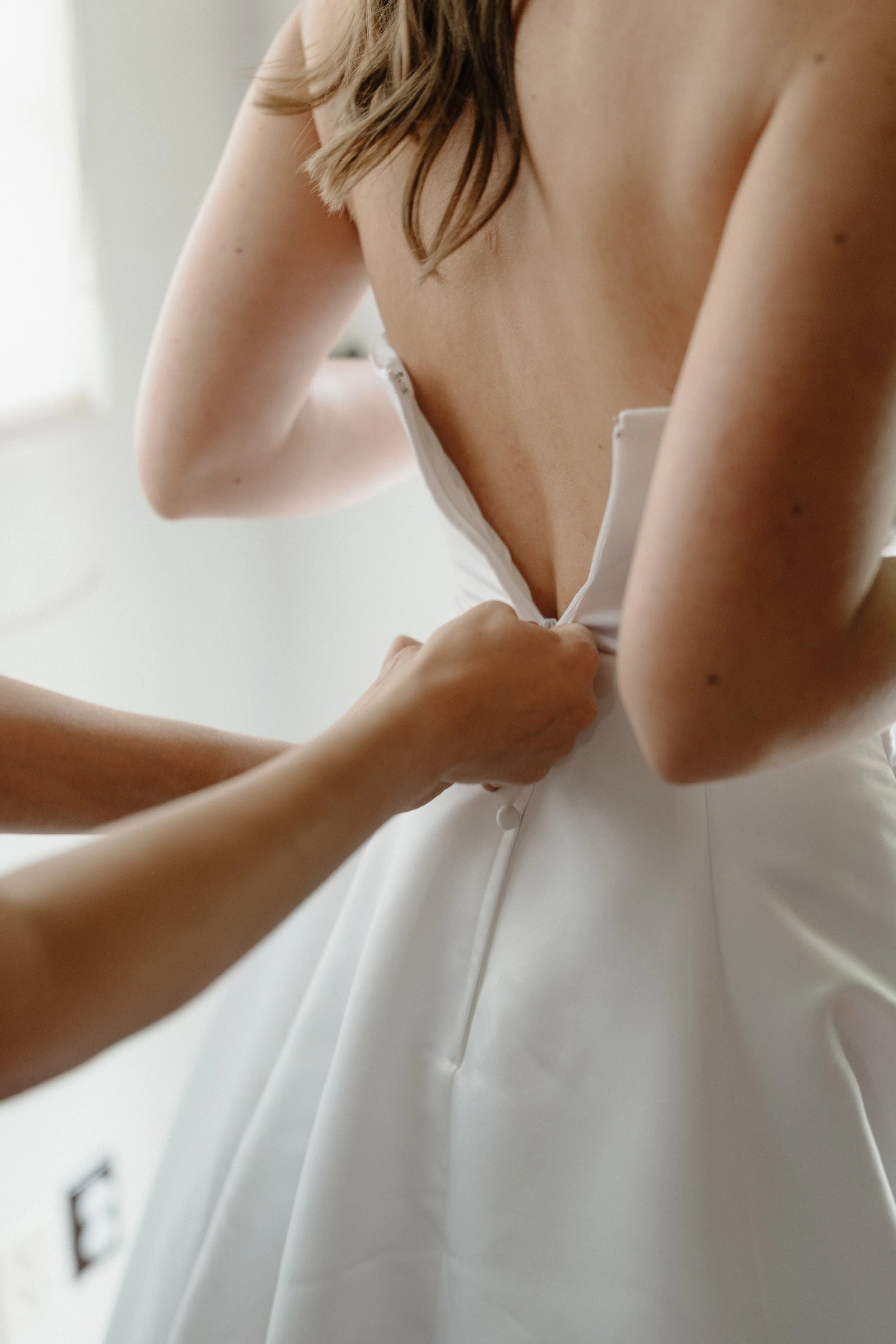 Close-up of a woman fastening the back of her white dress, showing her shoulder, arm, and part of her chest and neck.
