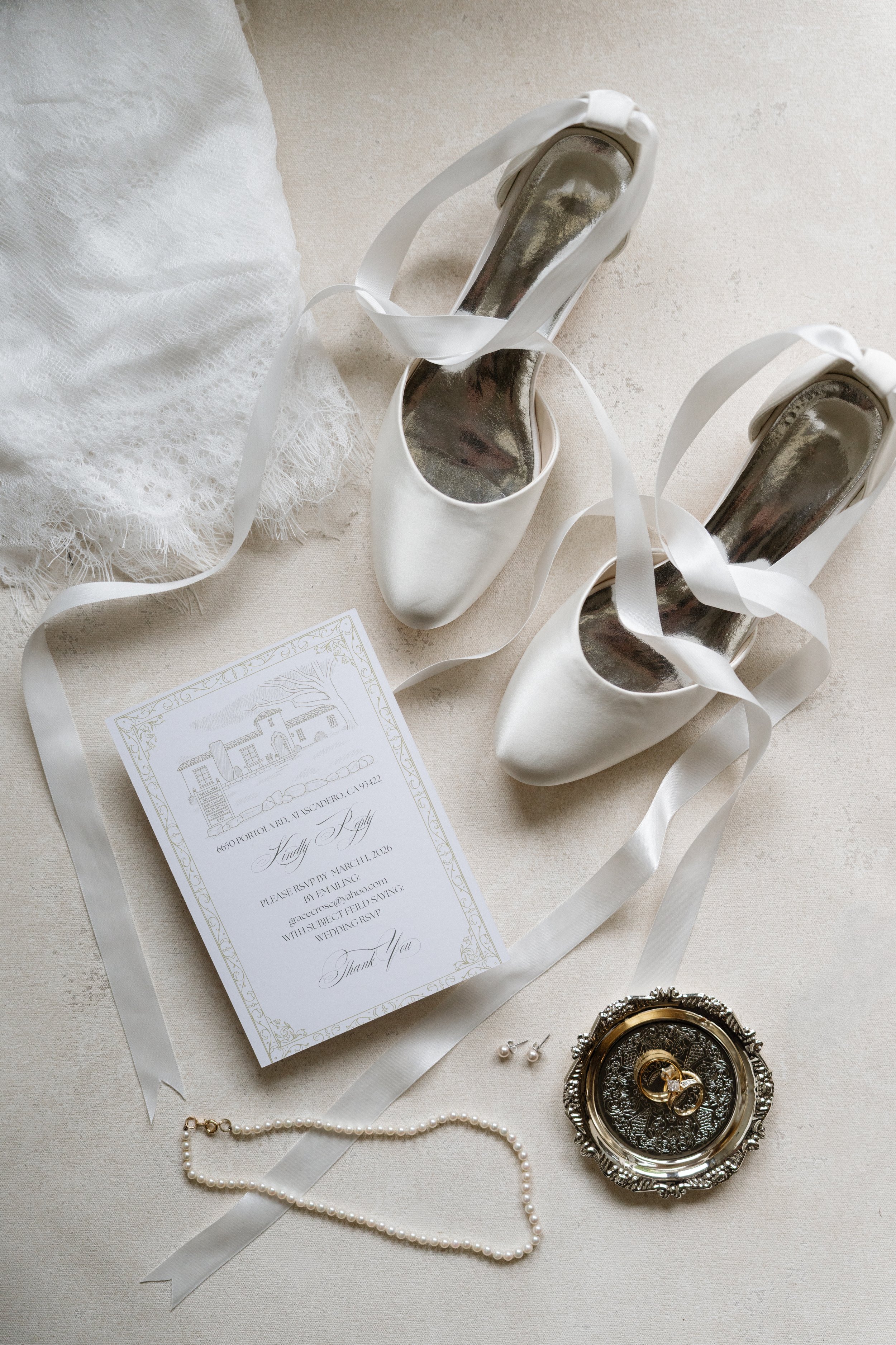 Silver high-heeled shoes with white ribbons, a wedding invitation, pearl necklace, and vintage pocket watch with jewelry.