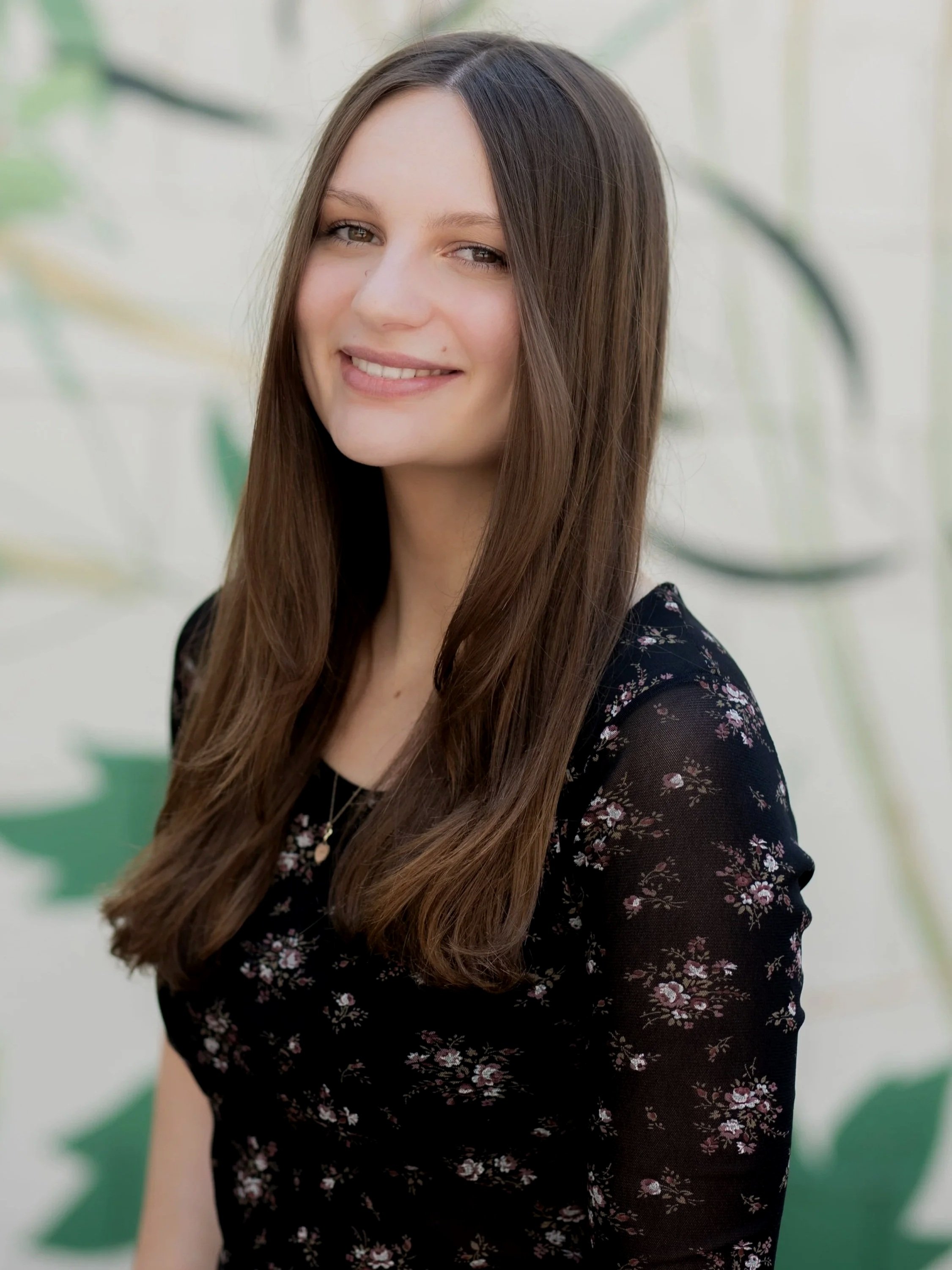 A young woman with long, wavy brown hair and light skin, smiling softly, wearing a black dress with pink flowers, standing in front of a light-colored, blurry background with some green leaves.