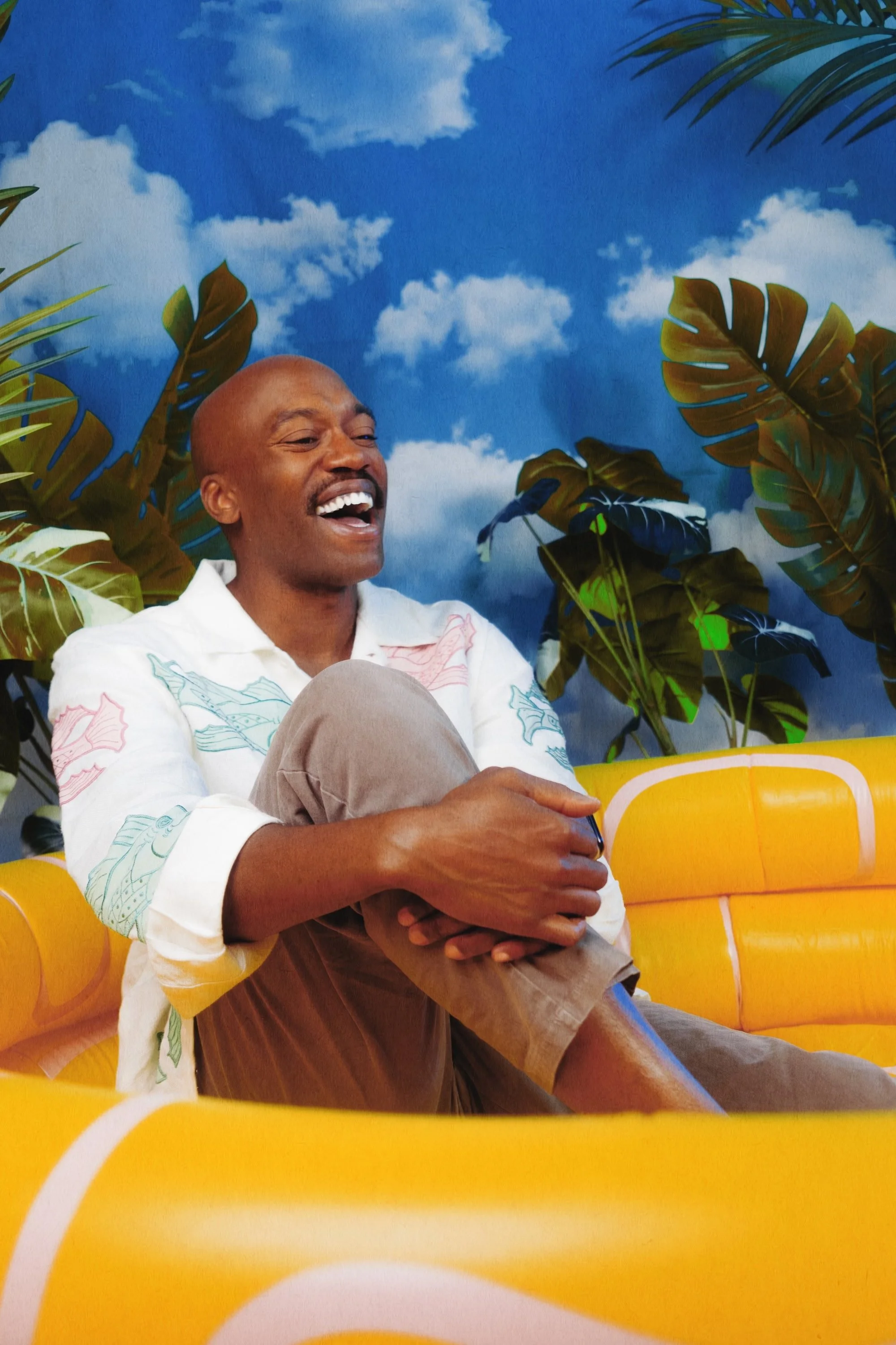 A man with a bald head and a mustache, wearing a white shirt with colorful patterns, laughing while sitting on a yellow bench with large green tropical leaves and a blue sky with white clouds painted background.