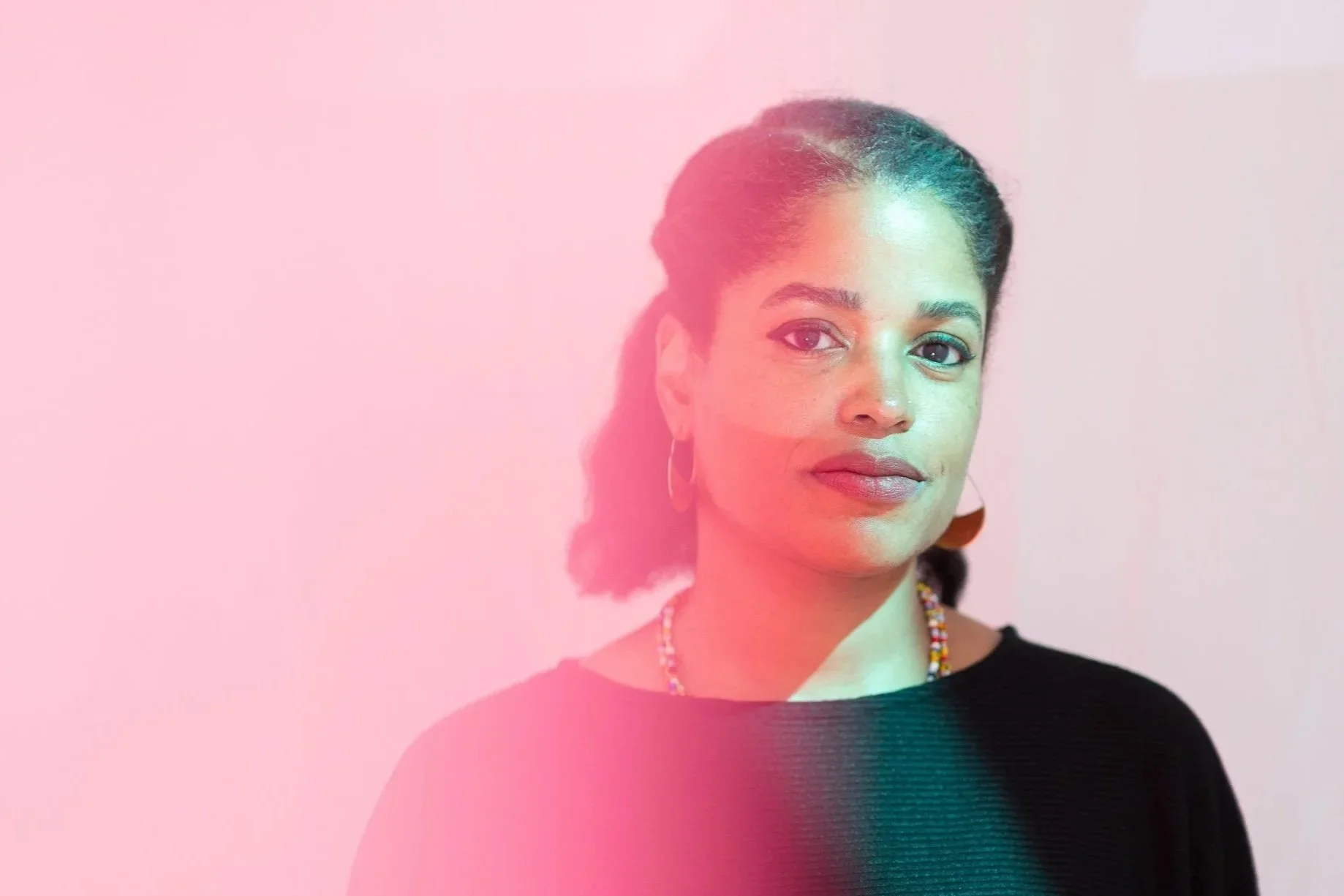 Portrait of a woman with braided hair, wearing earrings and a beaded necklace, against a pink background with a gradient effect.