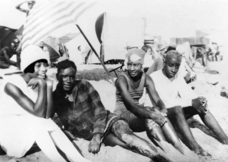 A vintage black and white photo from the 1950s of four Black people (two couples) sitting on the beach in Santa Monica, California with umbrellas in the background, wearing swimsuits and enjoying a sunny day.