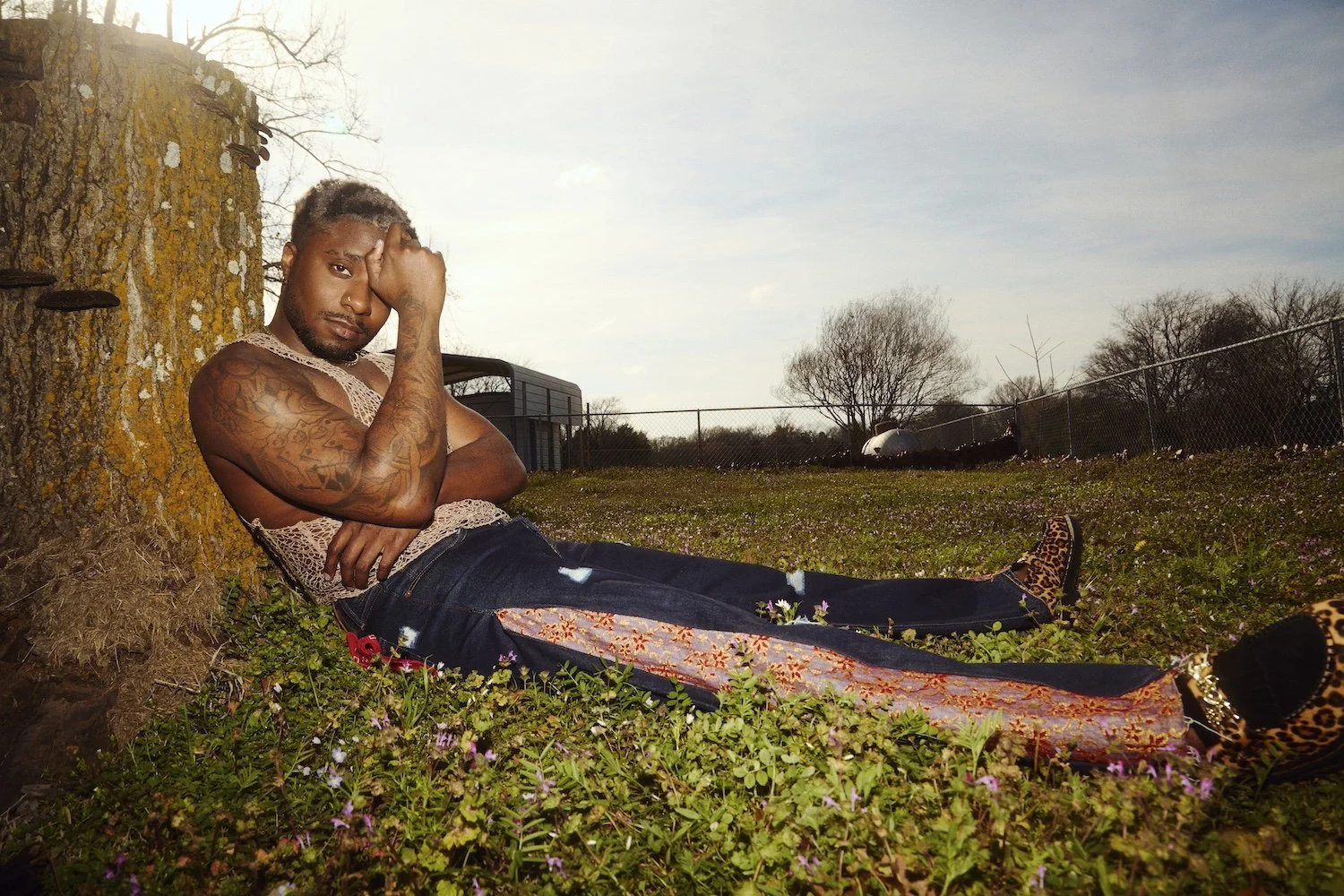 A Black man with tattoos and light-colored hair, wearing patterned pants and leopard print shoes, reclines in the grass next to a tree with a fence and trees in the background.