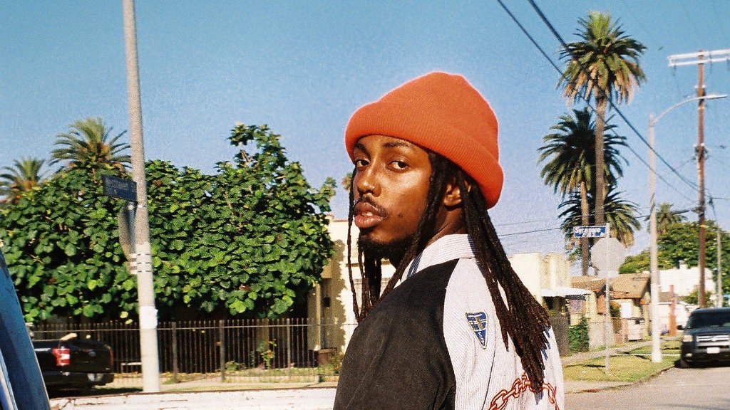 A man with long dreadlocks wearing an orange beanie and a black and white letterman jacket, standing outdoors on a sunny day with palm trees and houses in the background.