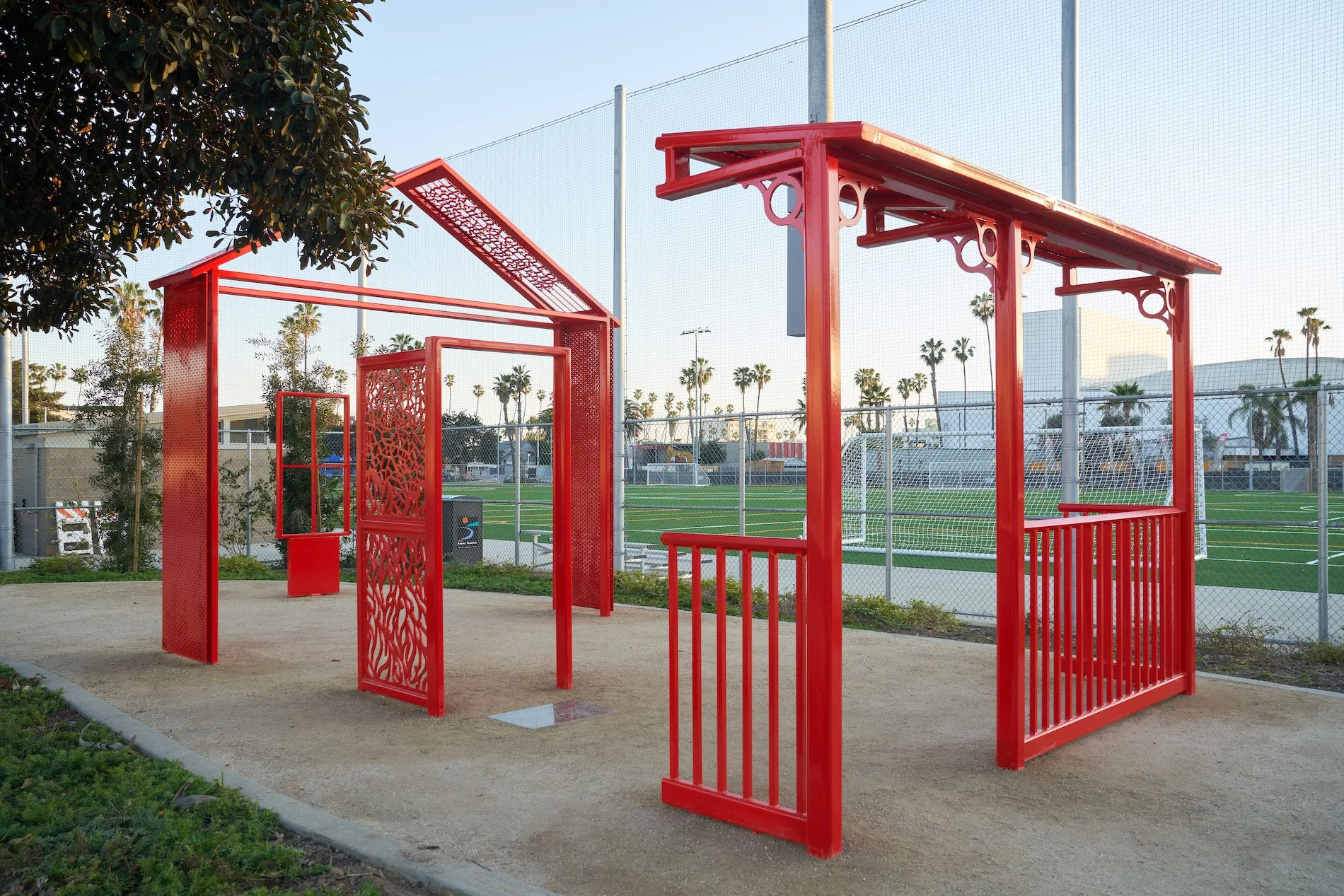 Red metal outdoor fitness structure with various bars and panels, situated on a dirt ground, near a chain-link fence with a soccer field and palm trees in the background, under a clear blue sky.