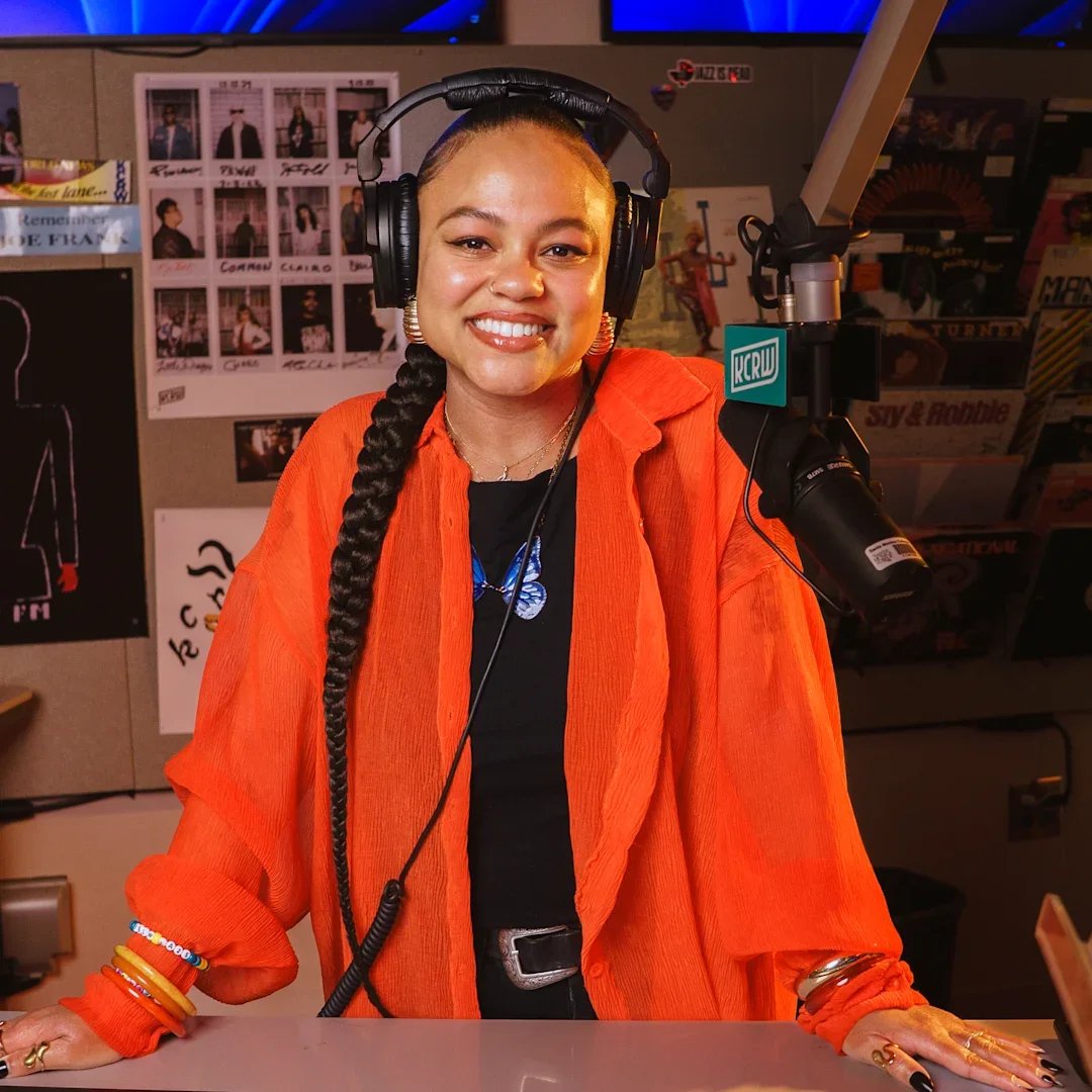 A woman with long braided hair, wearing headphones, an orange jacket over a black shirt, smiling, standing behind a desk at a radio station with a microphone.