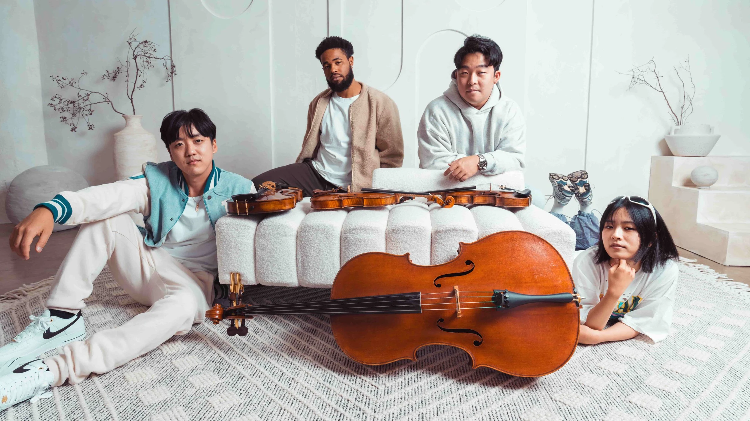 Four young adults in a minimalist room with white walls and decorative vases, posing with musical instruments including violas, violin, a cello, and a microphone, some sitting and some lying on the floor.