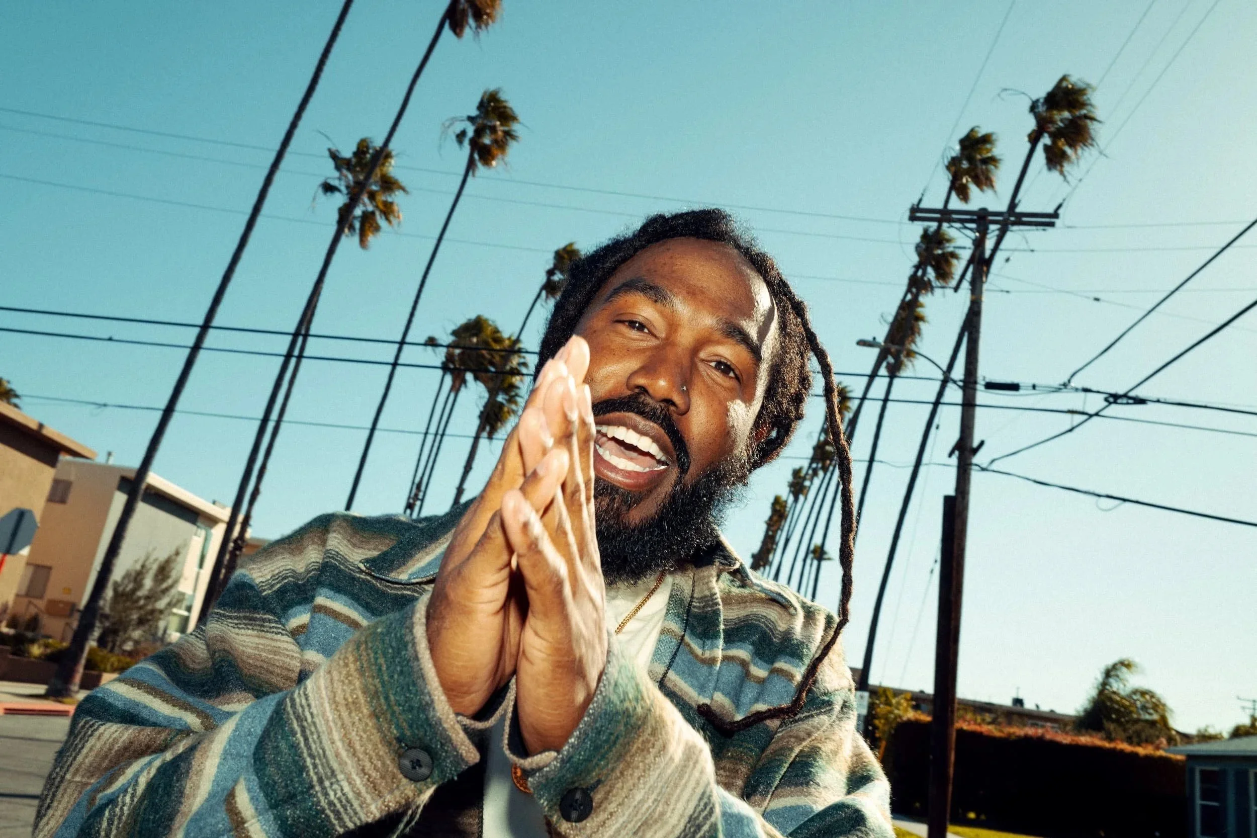 A man with dreadlocks and a beard smiling outdoors, wearing a plaid jacket, with power lines and palm trees in the background on a sunny day.