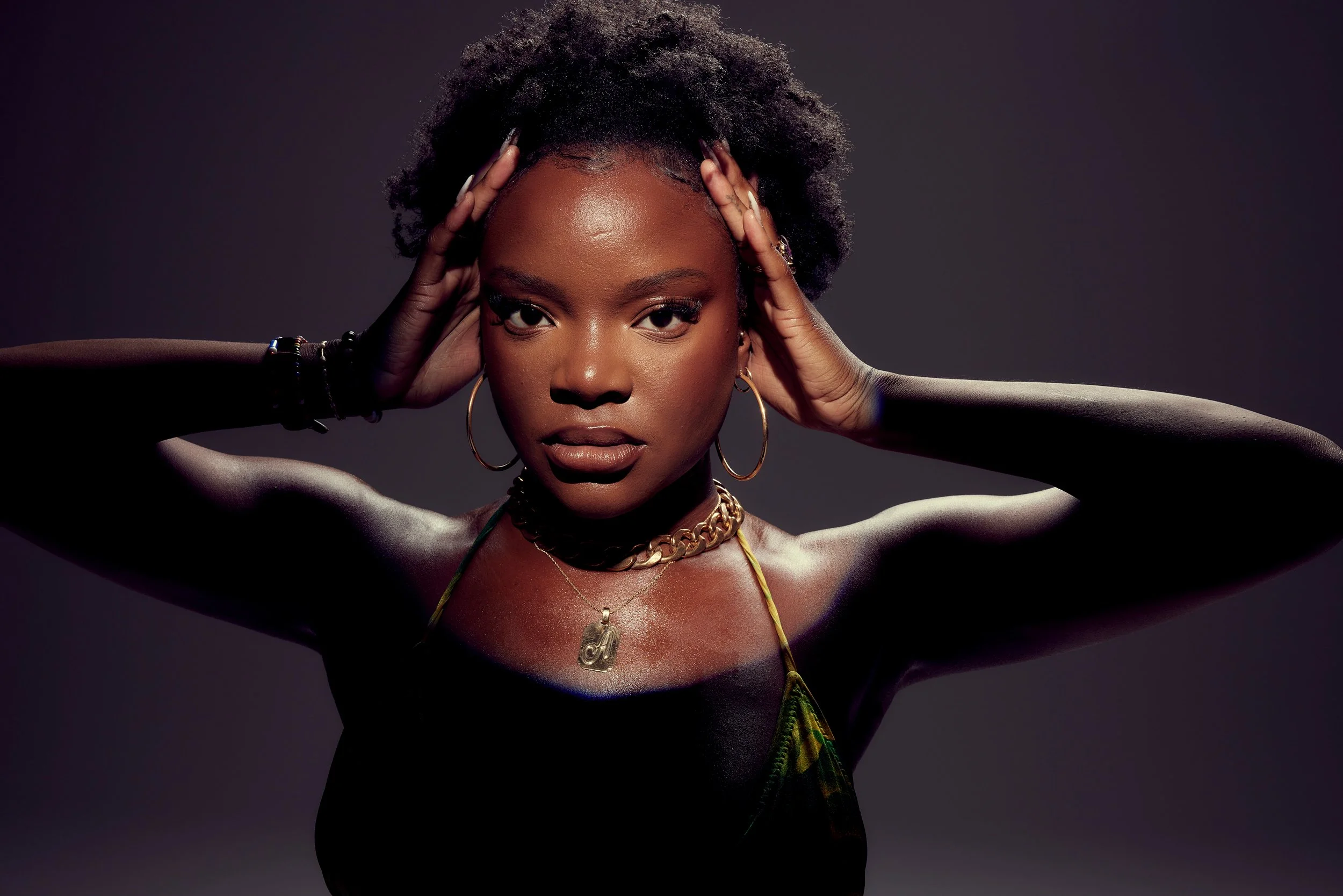 A woman with short natural curly hair and dark skin posing with hands on her head, wearing gold hoop earrings, necklaces, and a colorful top, against a dark background.
