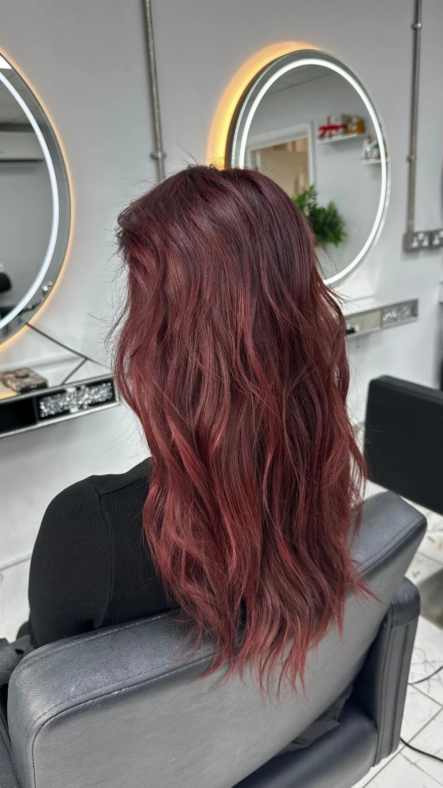 Back view of a woman with long, wavy, burgundy hair sitting in a salon chair with two illuminated round mirrors on the wall behind her.