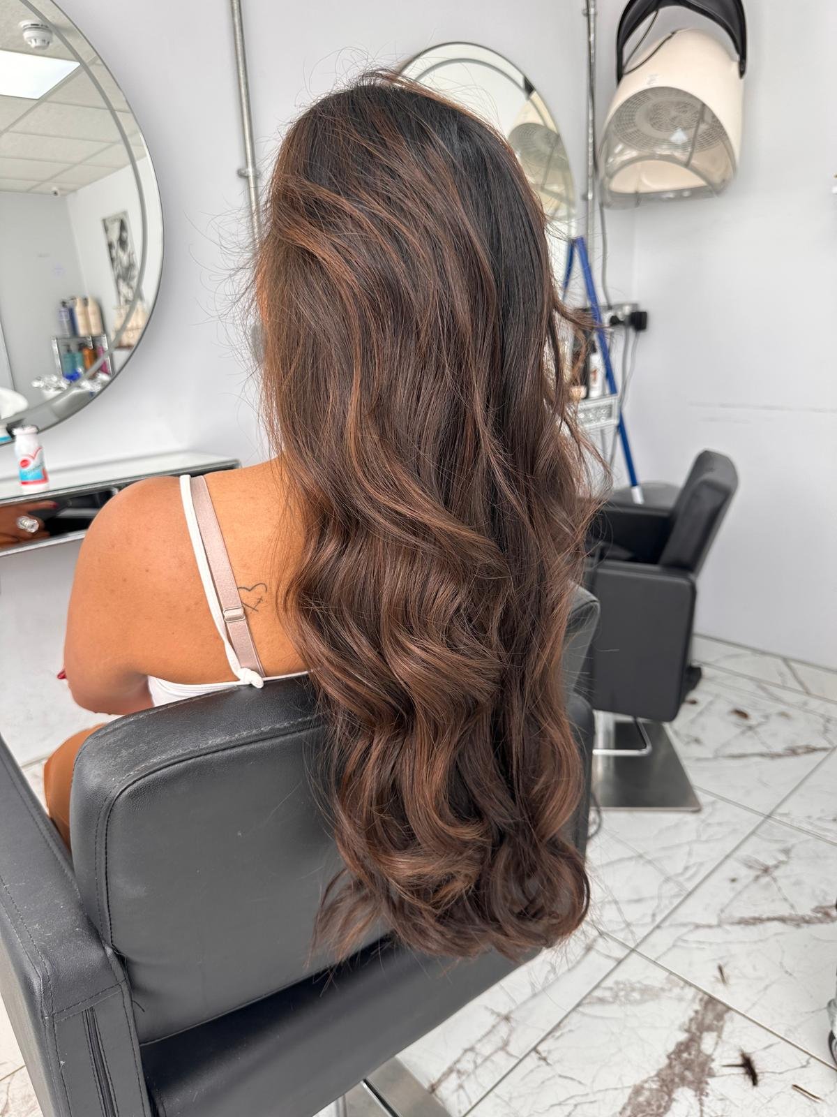A woman with wavy, brown hair sitting in a salon chair, facing away from the camera, with a mirror and salon tools visible in the background.