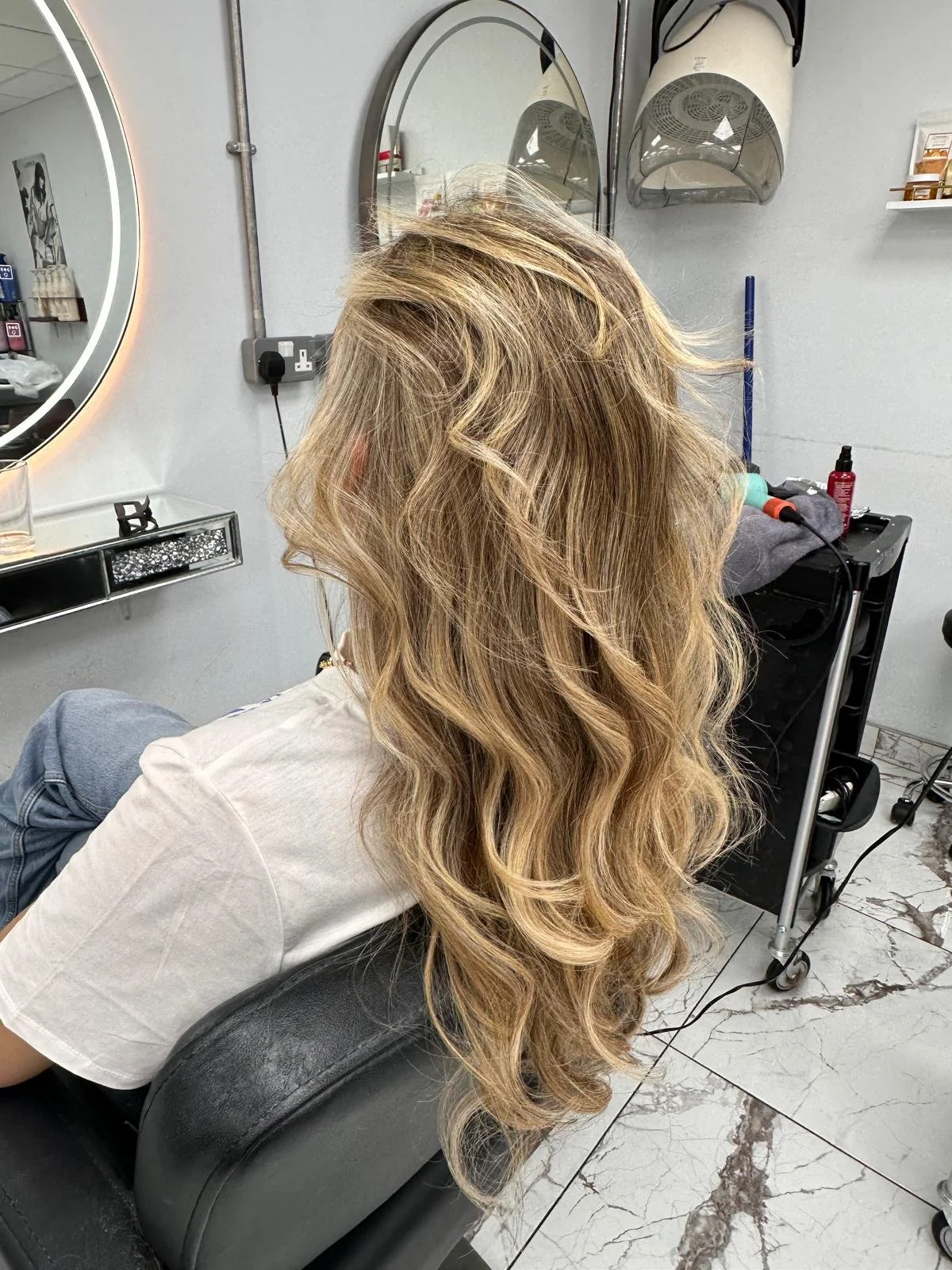 A woman with long, wavy blonde hair sitting in a salon chair with her back facing the mirror, getting her hair styled.