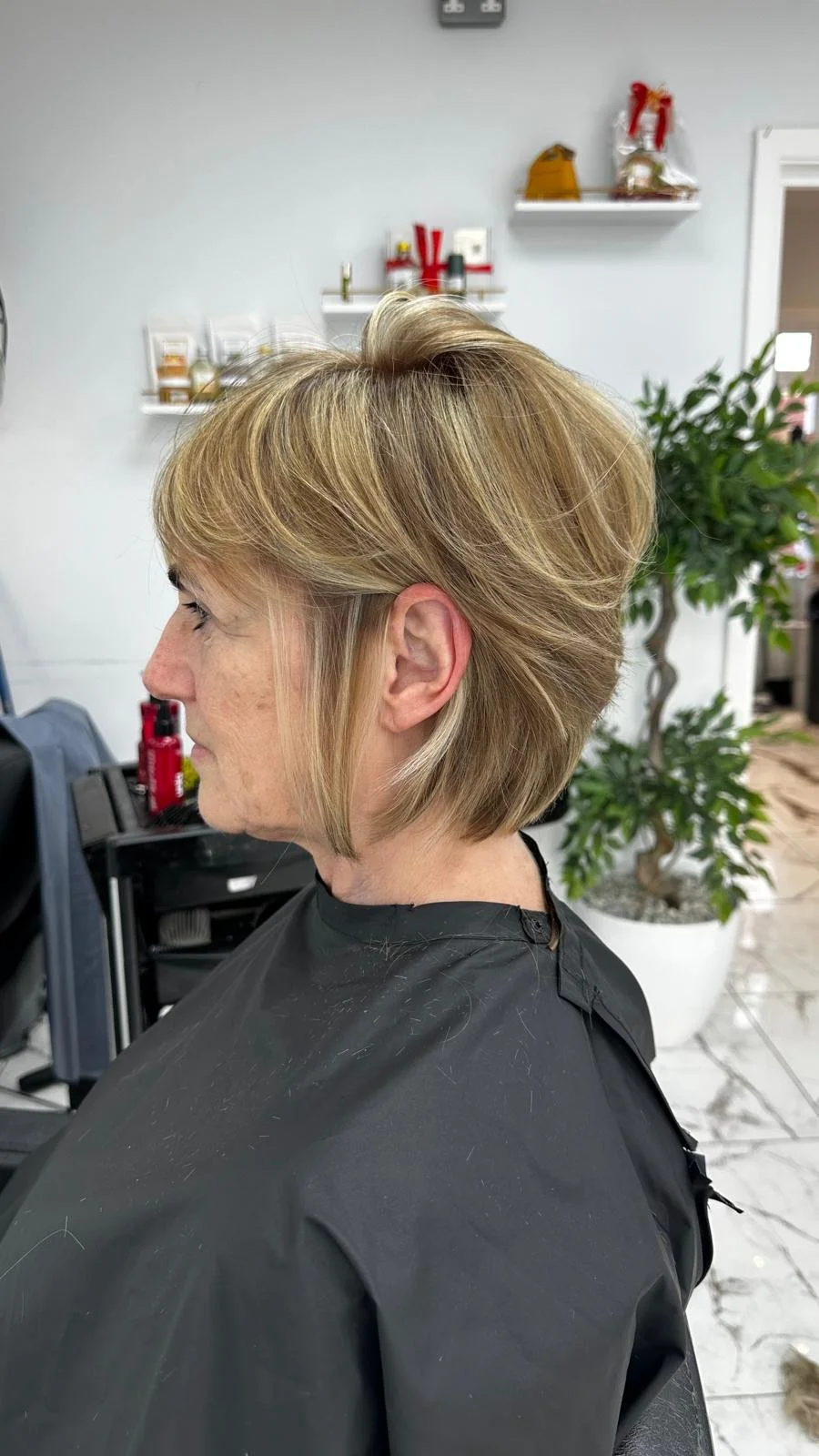 Side view of an older woman with short, blonde hair in a professional hair salon, sitting in a black cape, with shelves and a potted plant in the background.