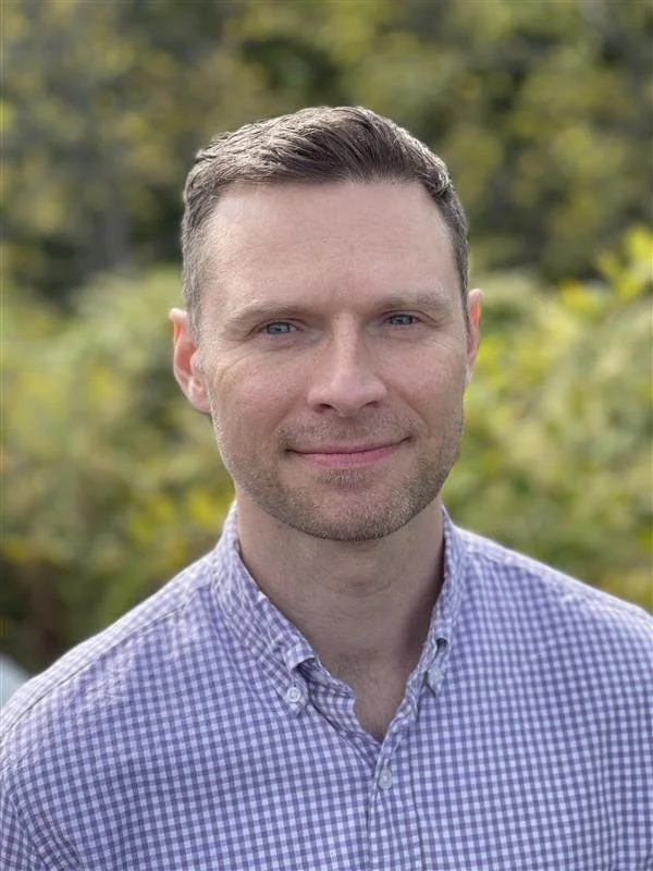 A man with short brown hair and blue eyes wearing a purple and white checkered button-up shirt outdoors with blurred green trees in the background.