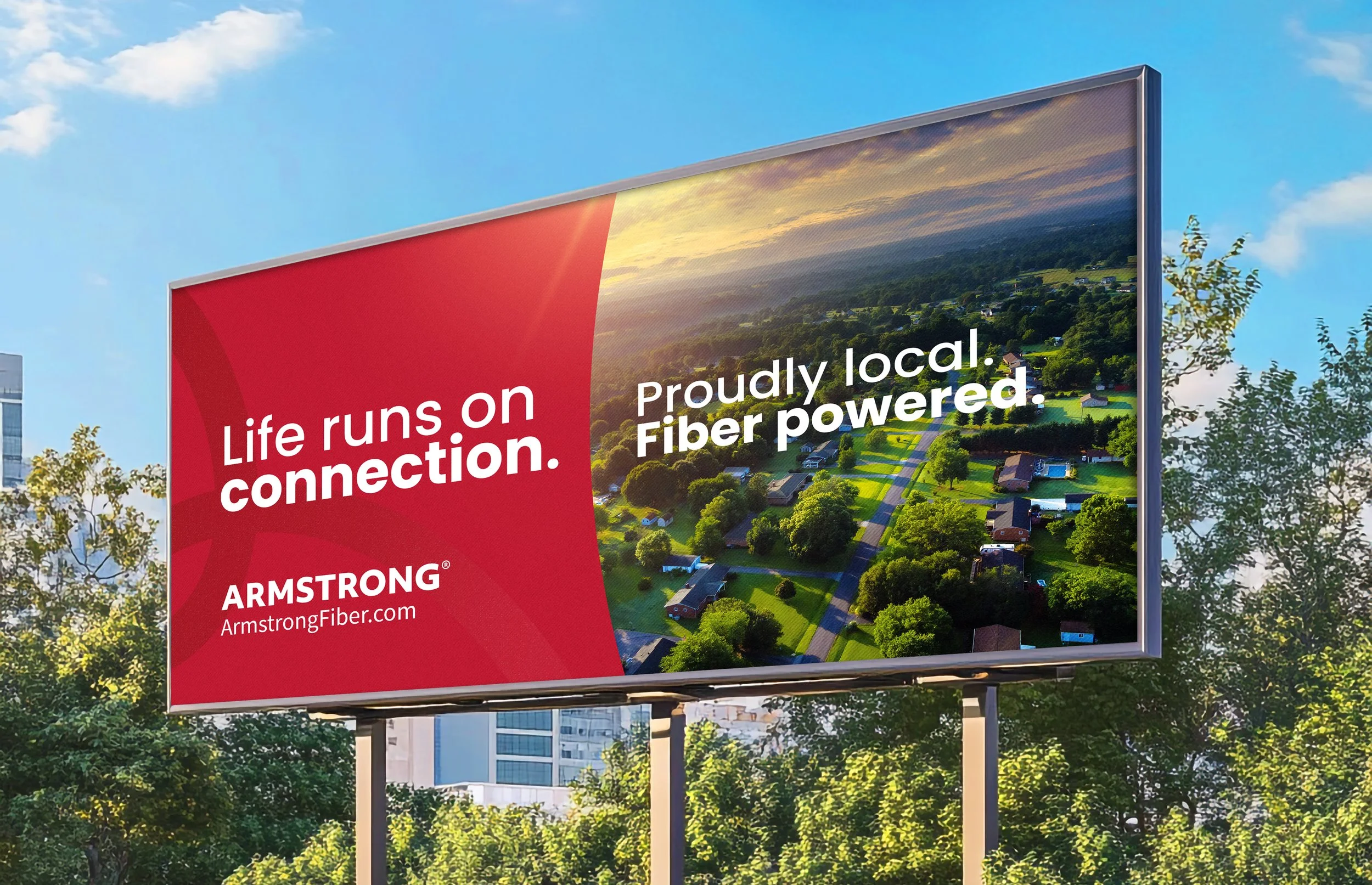 Billboard with a red section on the left and a scenic aerial view of a neighborhood with houses, trees, and roads on the right. The billboard has text promoting fiber internet service, with the phrases "Life runs on connection" and "Proudly local. Fiber powered."