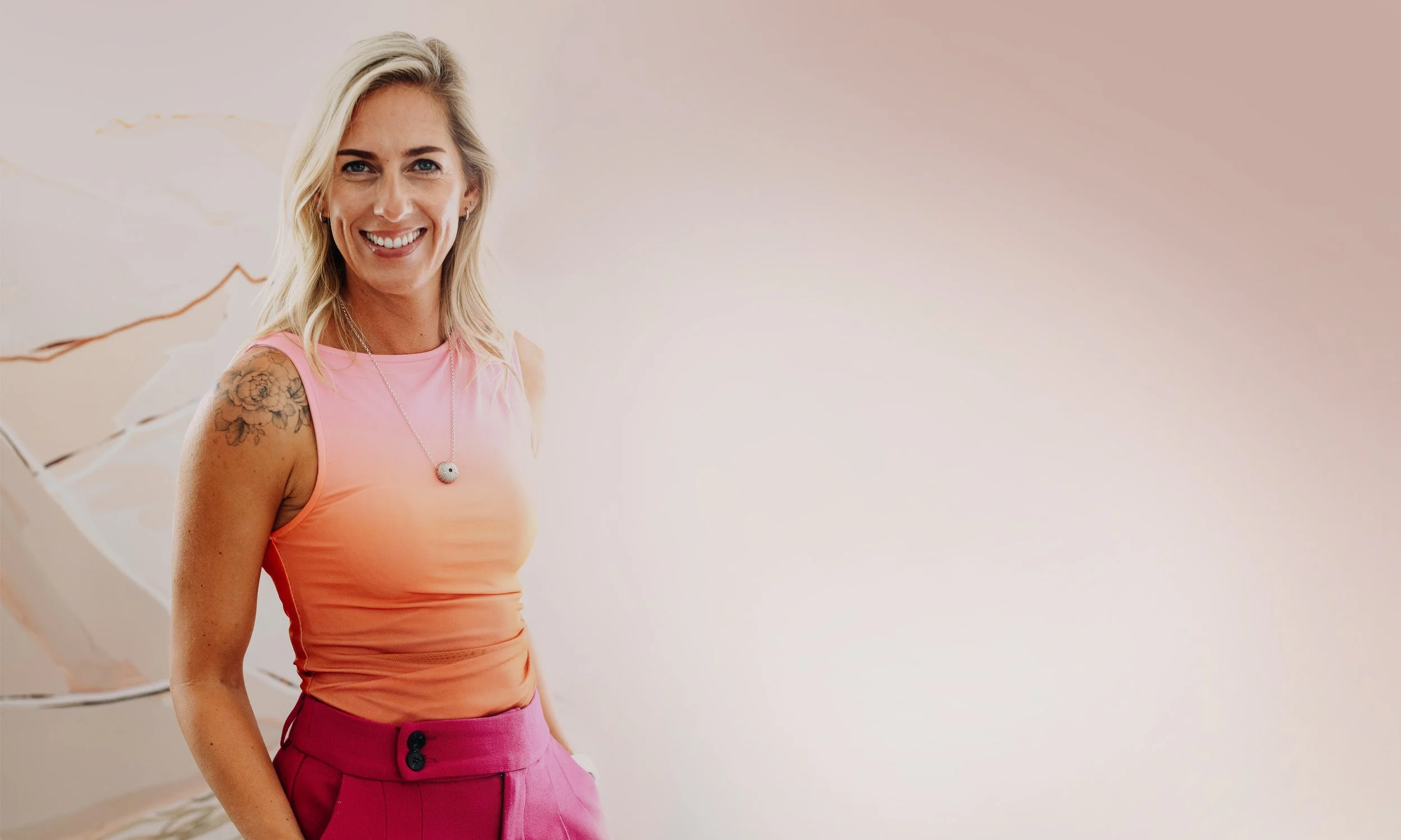 A smiling woman with blonde hair, tattoos on her arm, wearing a pink sleeveless top and magenta pants, standing indoors against a plain wall with a decorative picture frame in the background.