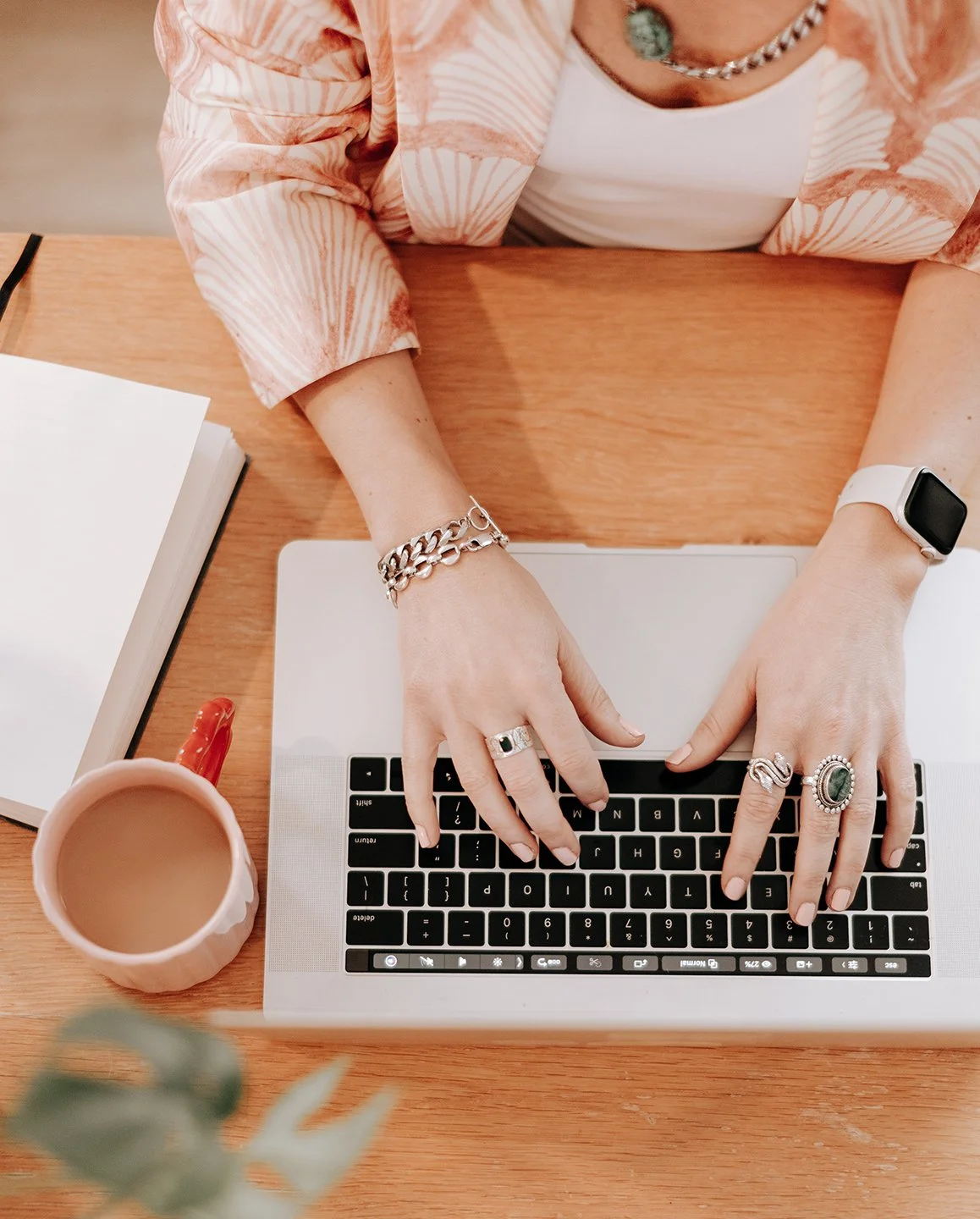 A person working on a laptop at a wooden desk with jewelry, rings, bracelets, and a smart watch, a mug, an open notebook, and a keychain nearby.