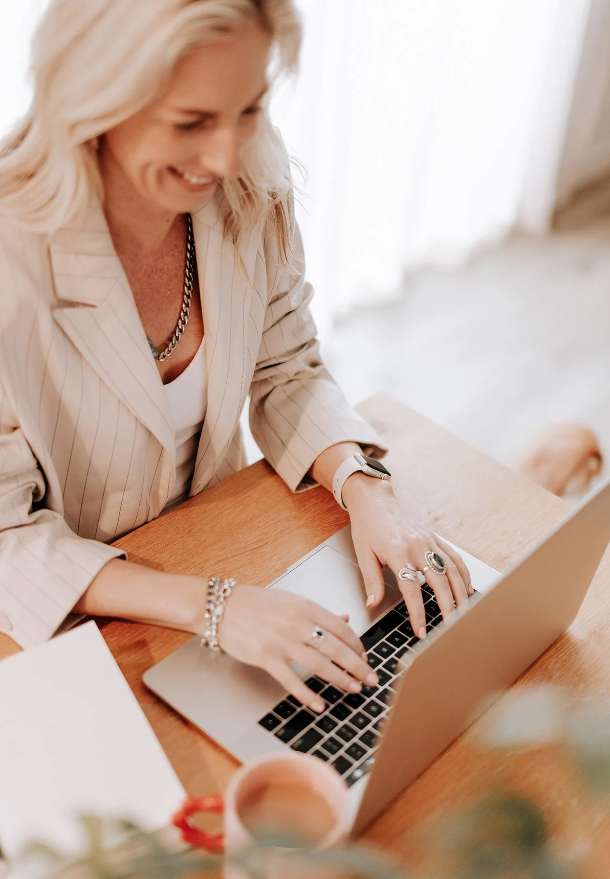 A woman with blonde hair, wearing a beige pinstripe blazer, working on a laptop at a wooden table, smiling and wearing rings and jewelry.