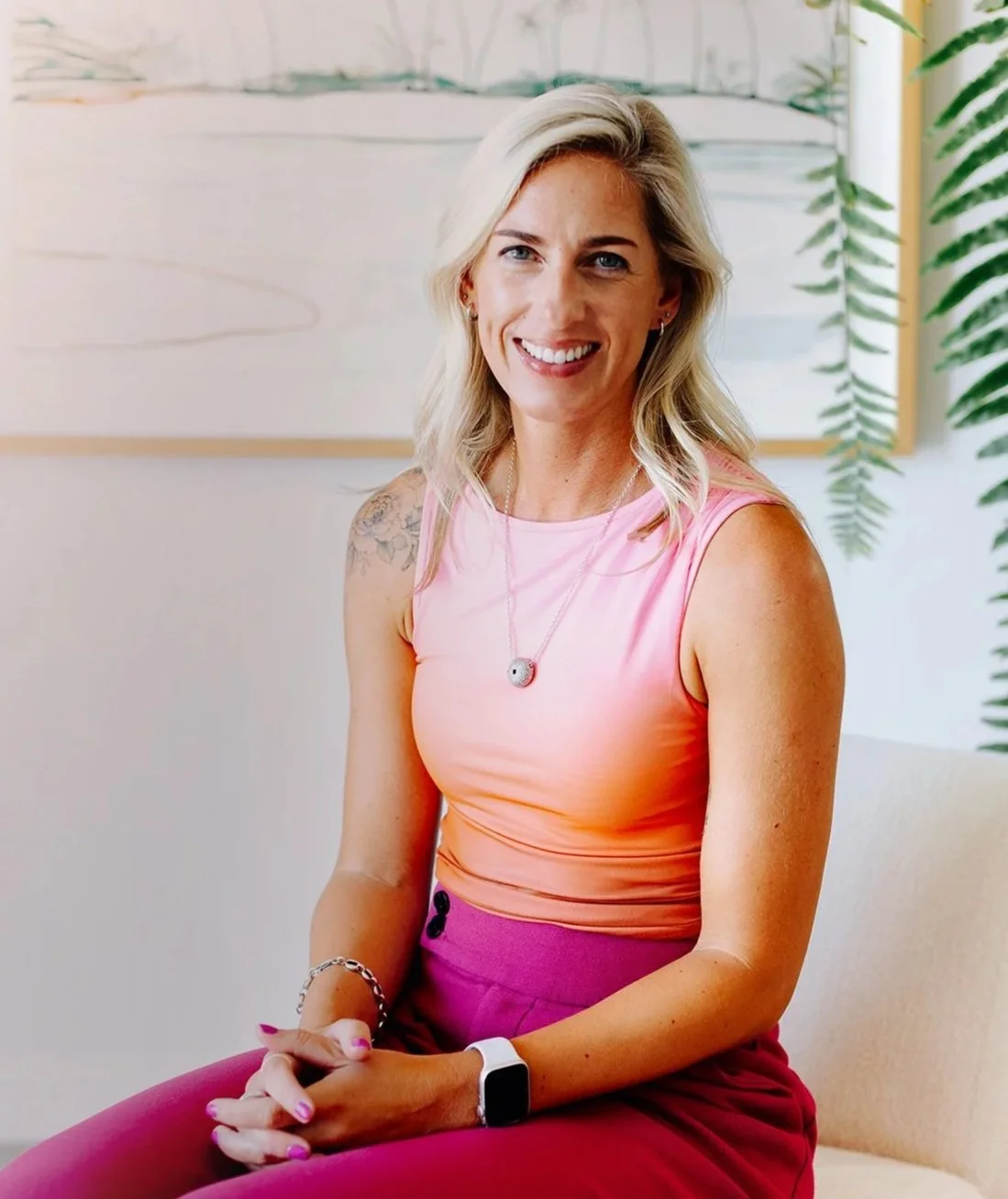 A woman with blonde hair smiling, wearing a pink sleeveless top, pink pants, a smartwatch, and a necklace, sitting indoors with green plants and artwork in the background.