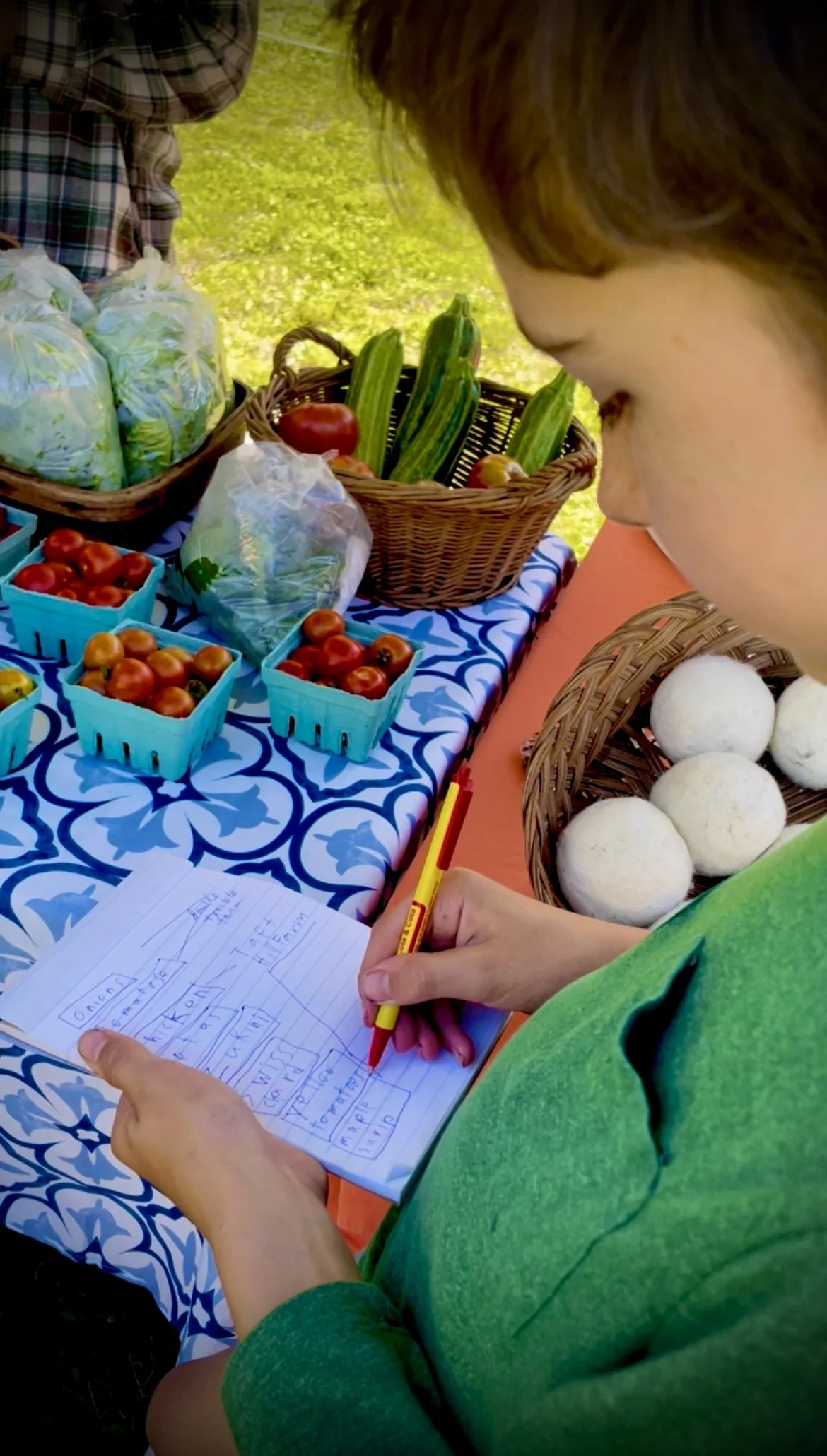 Child writing notes at a table with fresh vegetables and fruits, including tomatoes, cucumbers, and mushrooms, at an outdoor market.