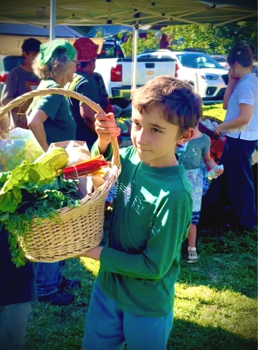 A young boy holding a basket of vegetables at an outdoor market or fair with several people and parked cars in the background.