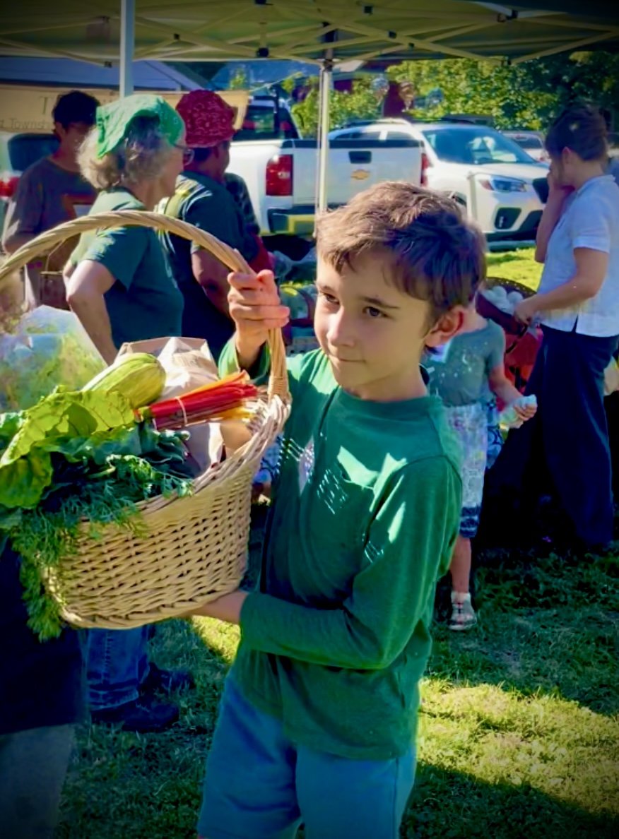 A young boy holding a basket of fresh vegetables at an outdoor market with other people shopping and vehicles in the background.