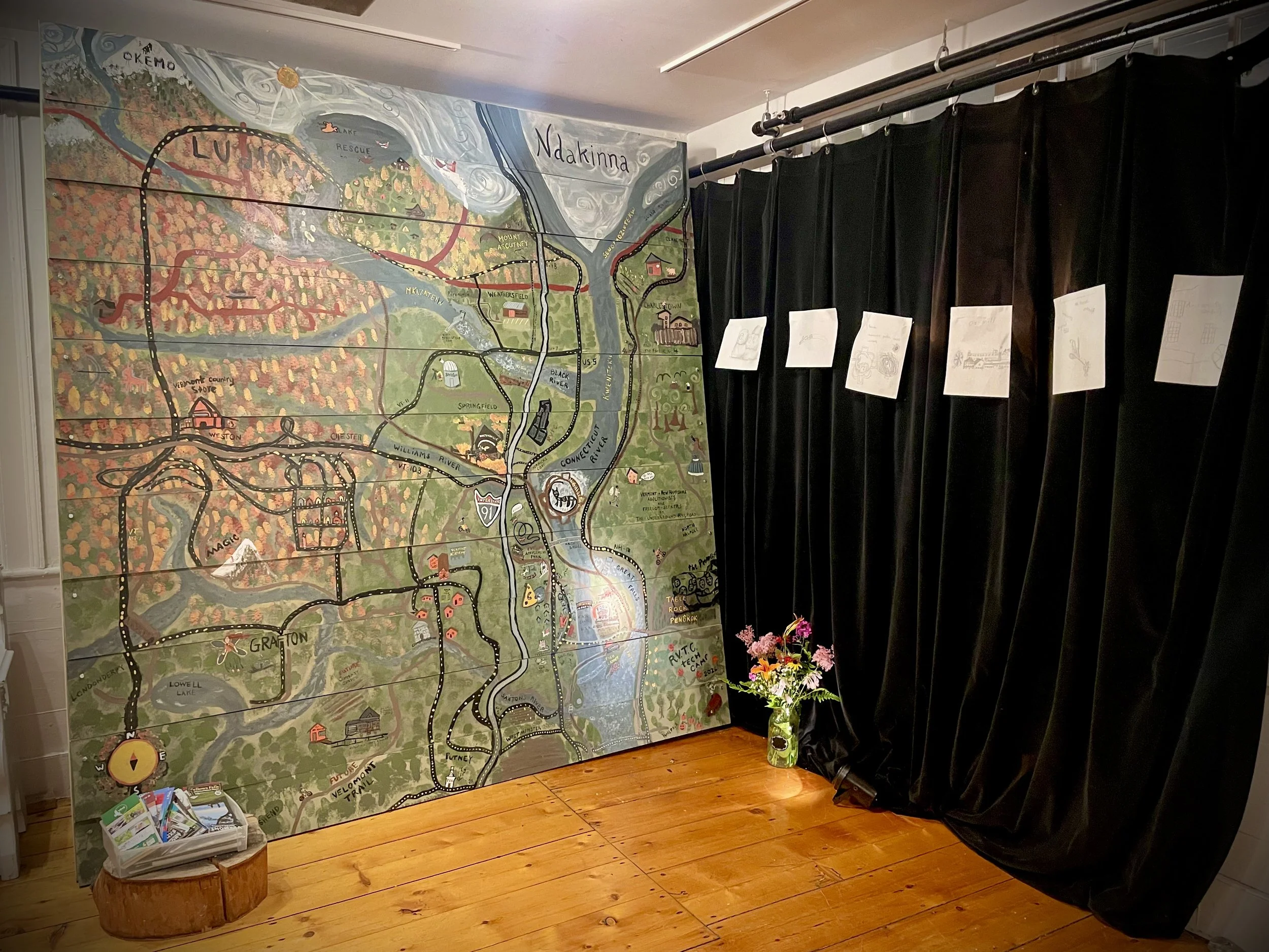 A large illustrated map of Vermont and surrounding areas displayed on a wooden wall, with black curtains hanging on the right side, and a small wooden stool with a container of brochures and a green plant with pink and orange flowers in front.